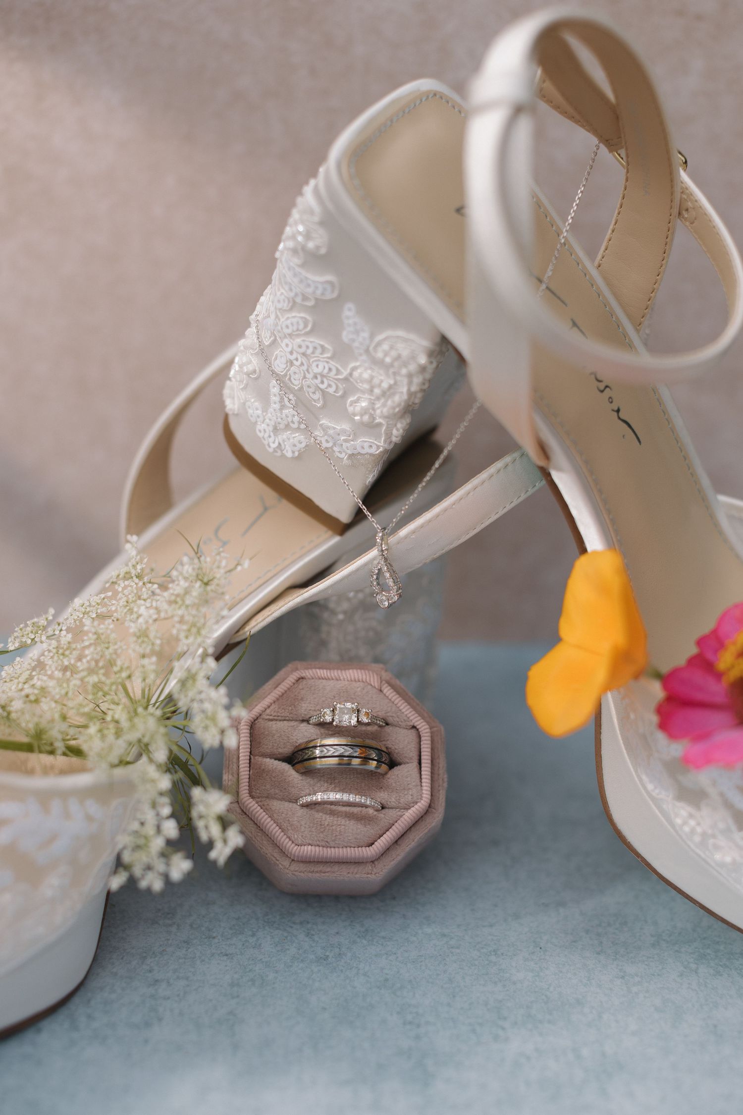 White lace bridal heels displayed with a pink velvet ring box holding diamond wedding bands and floral accents.