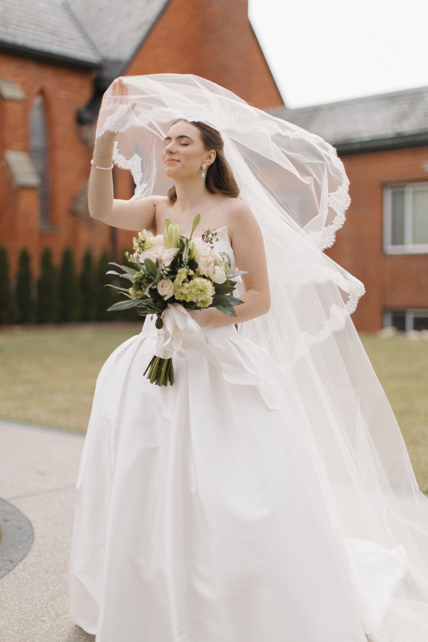 A bride in a flowing white dress holds a bouquet while her veil catches the breeze outdoors.