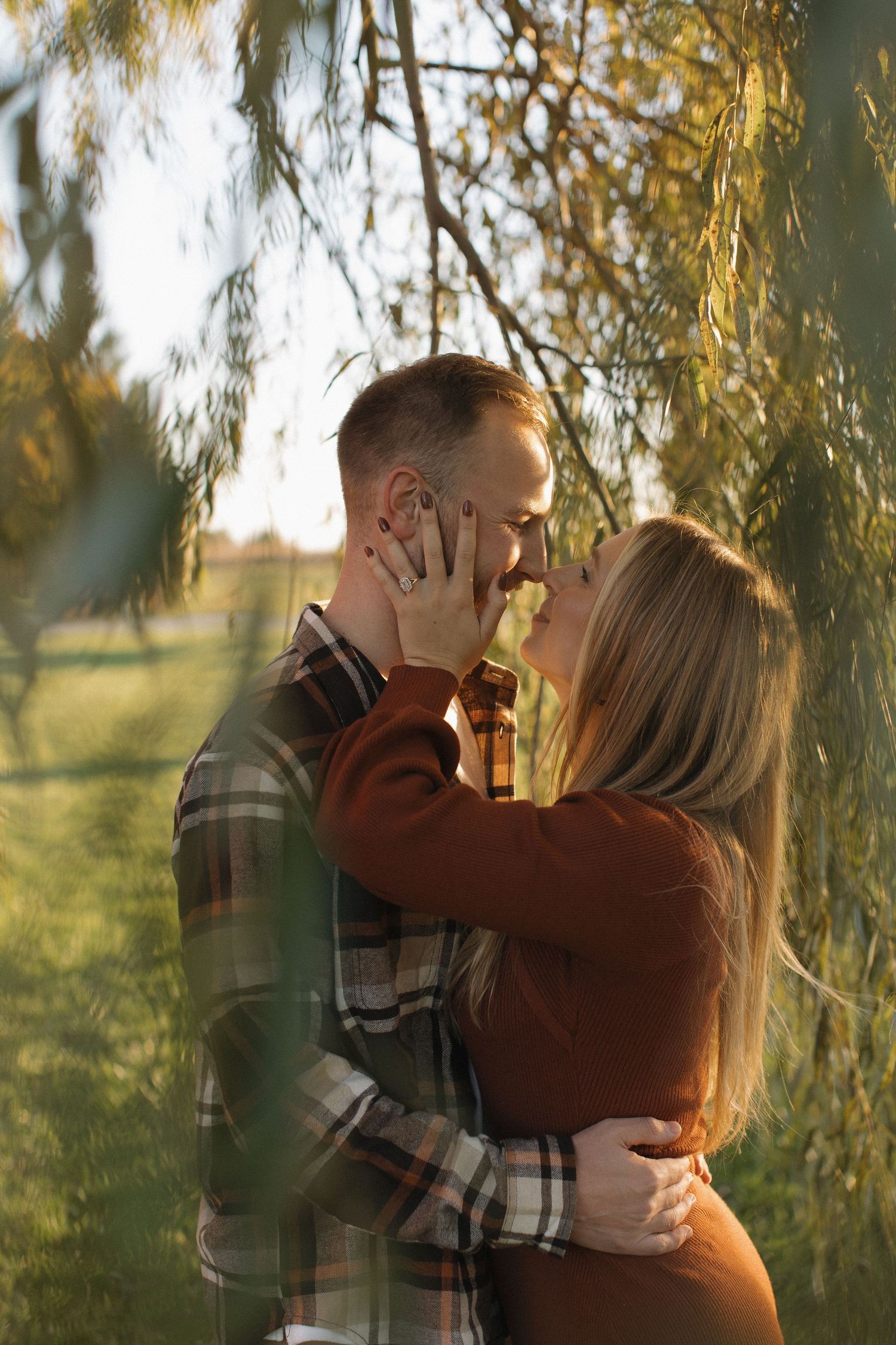 A romantic couple embraces under willow branches during a golden sunset photo session.