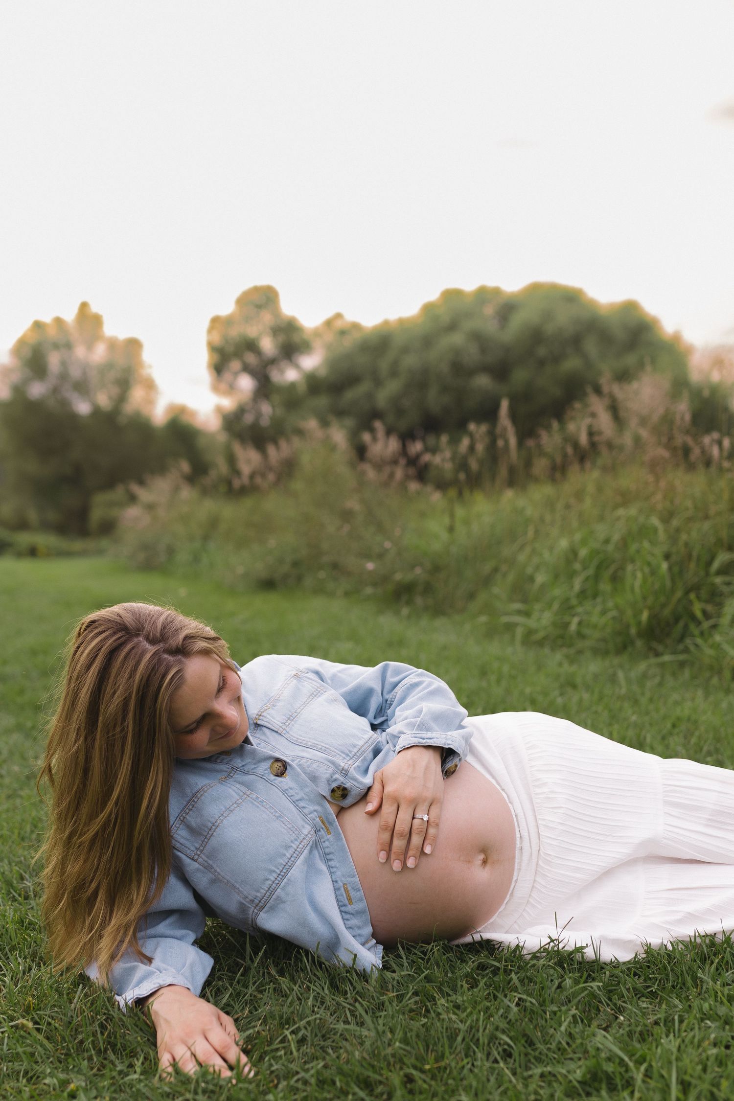 Pregnant woman lying on grass in white dress and denim jacket, tenderly touching her baby bump outdoors.