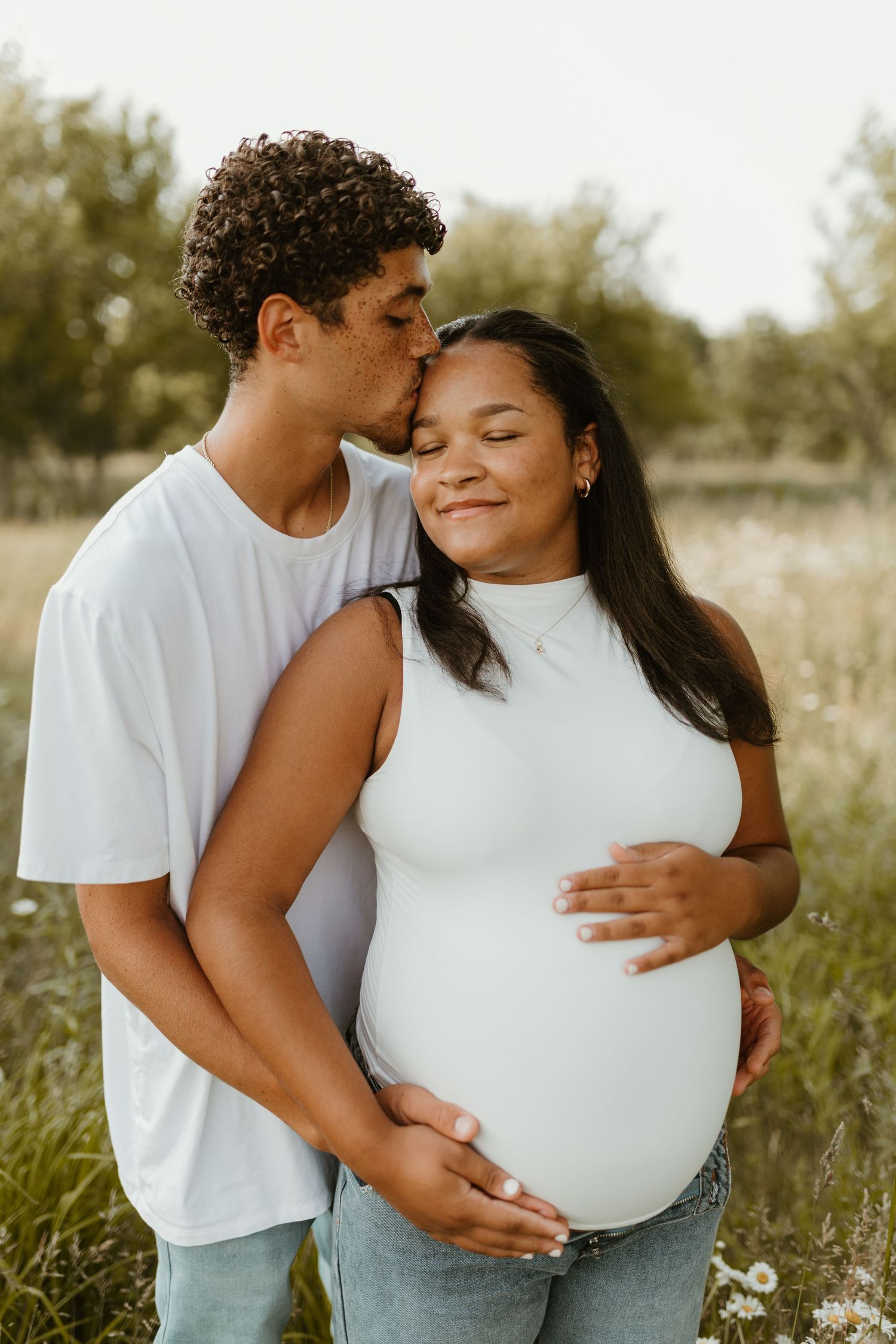 A tender maternity photoshoot in a sunlit field with a couple embracing in casual white and denim outfits.