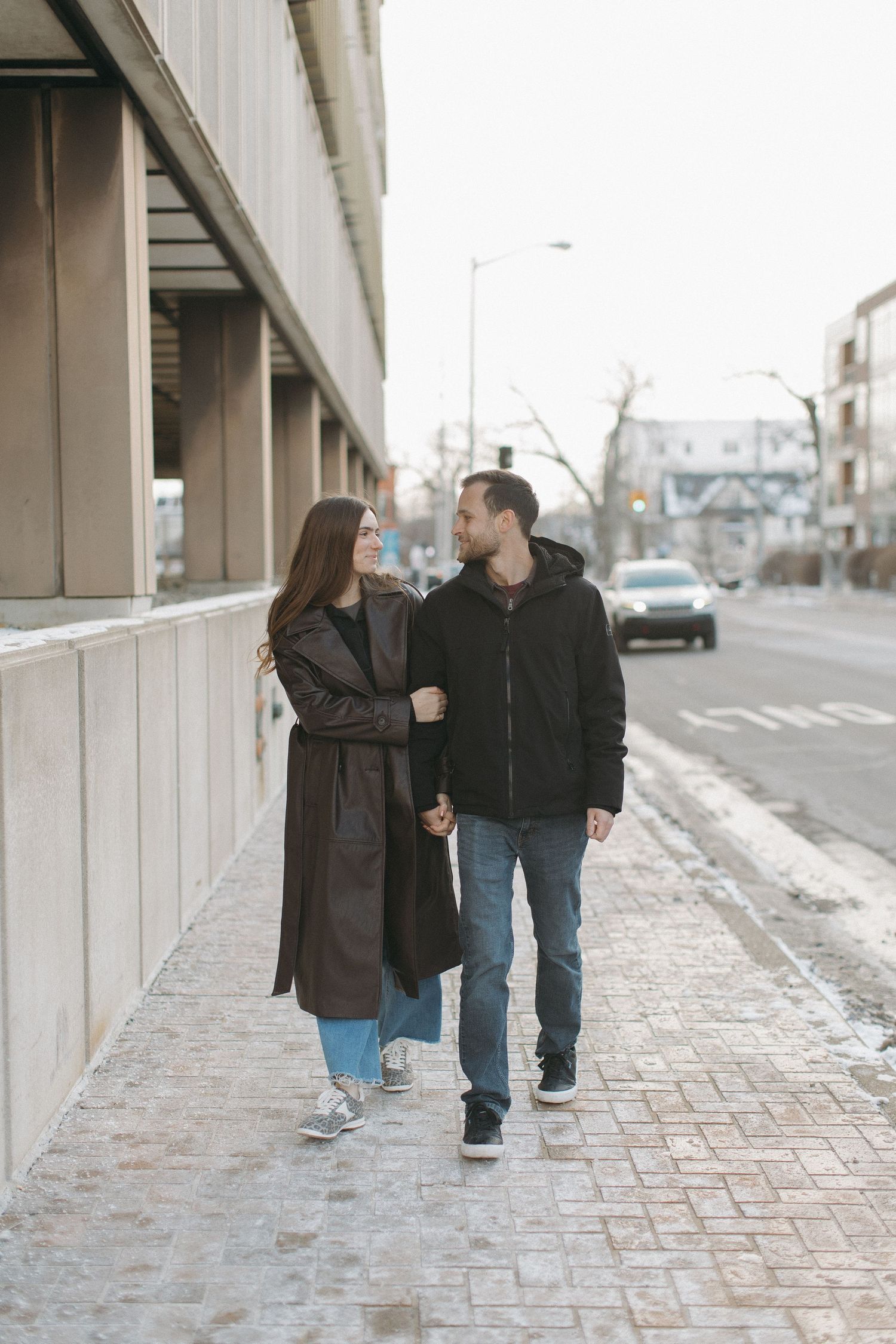 Casual couple walking along urban sidewalk during winter engagement session.