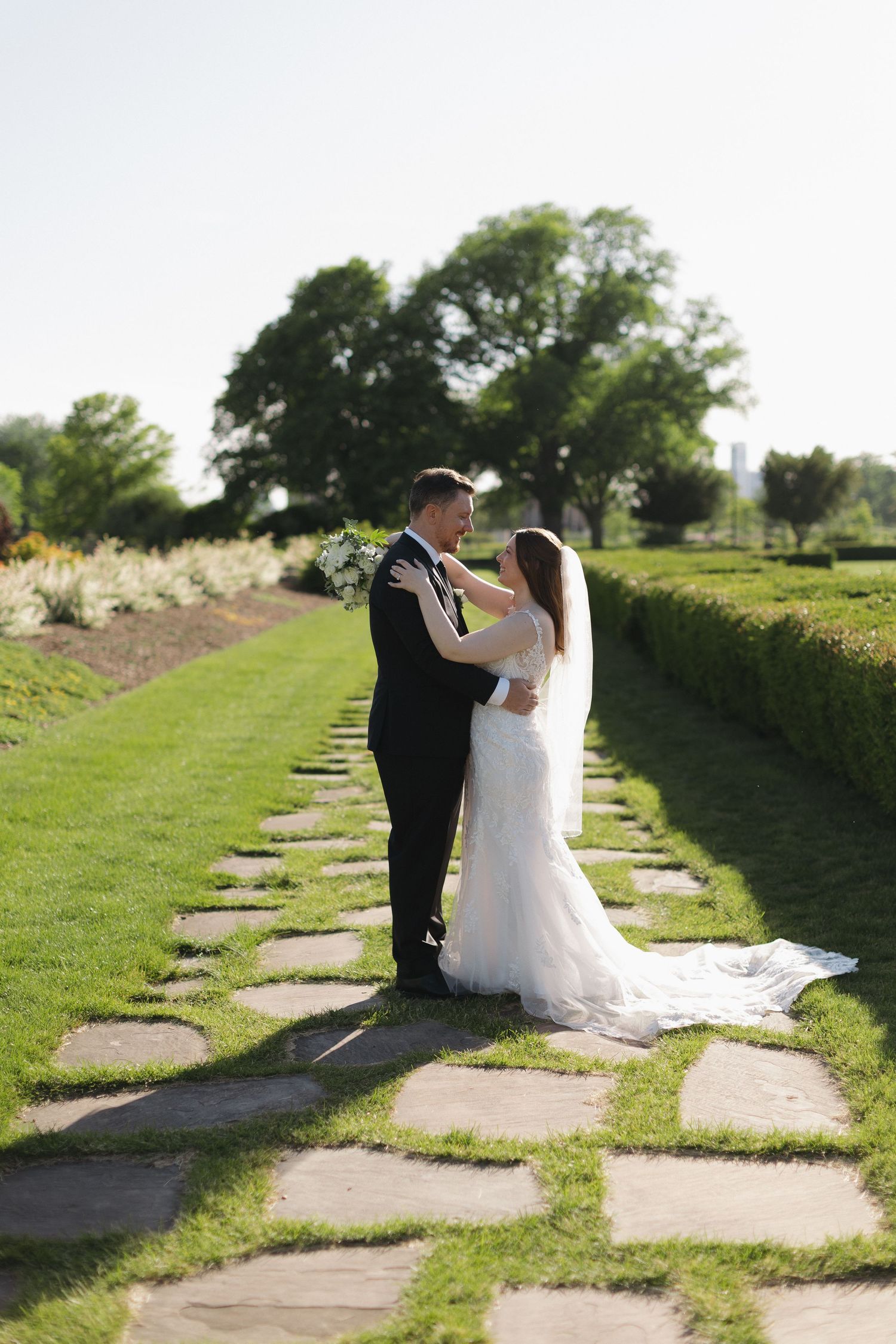 Bride and groom embrace on stone path in lush garden, bride in white gown with veil, groom in black suit.