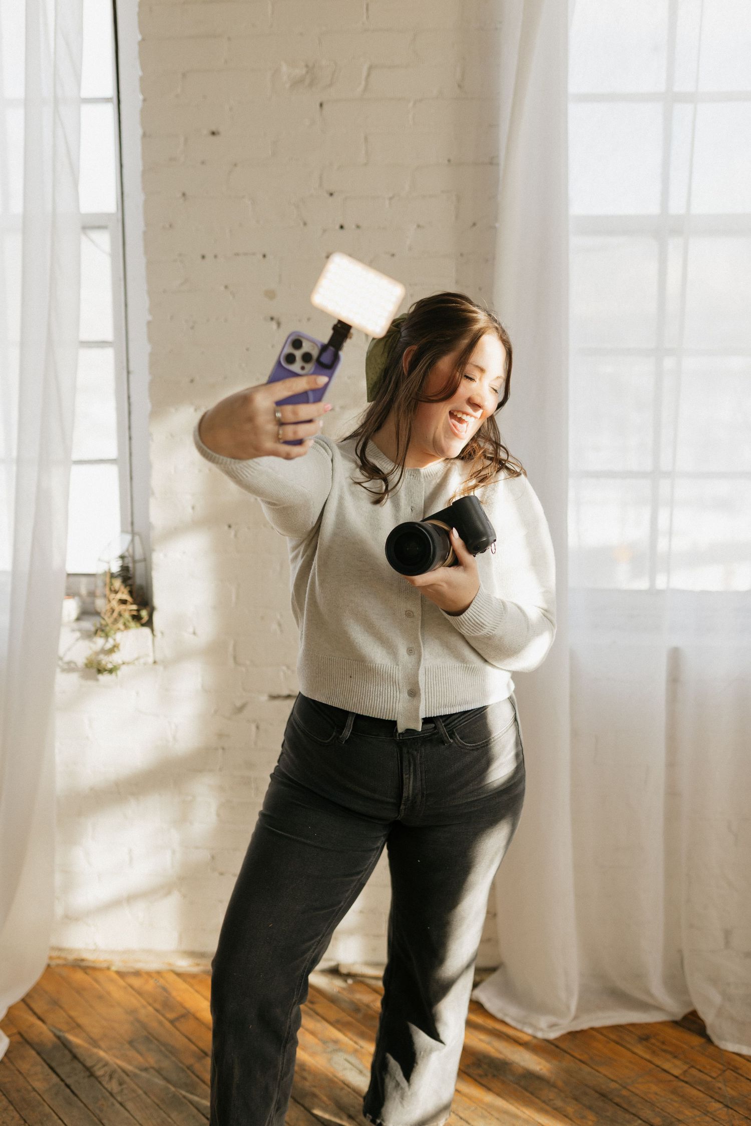 Woman in light sweater dancing and taking photos in sunlit studio.