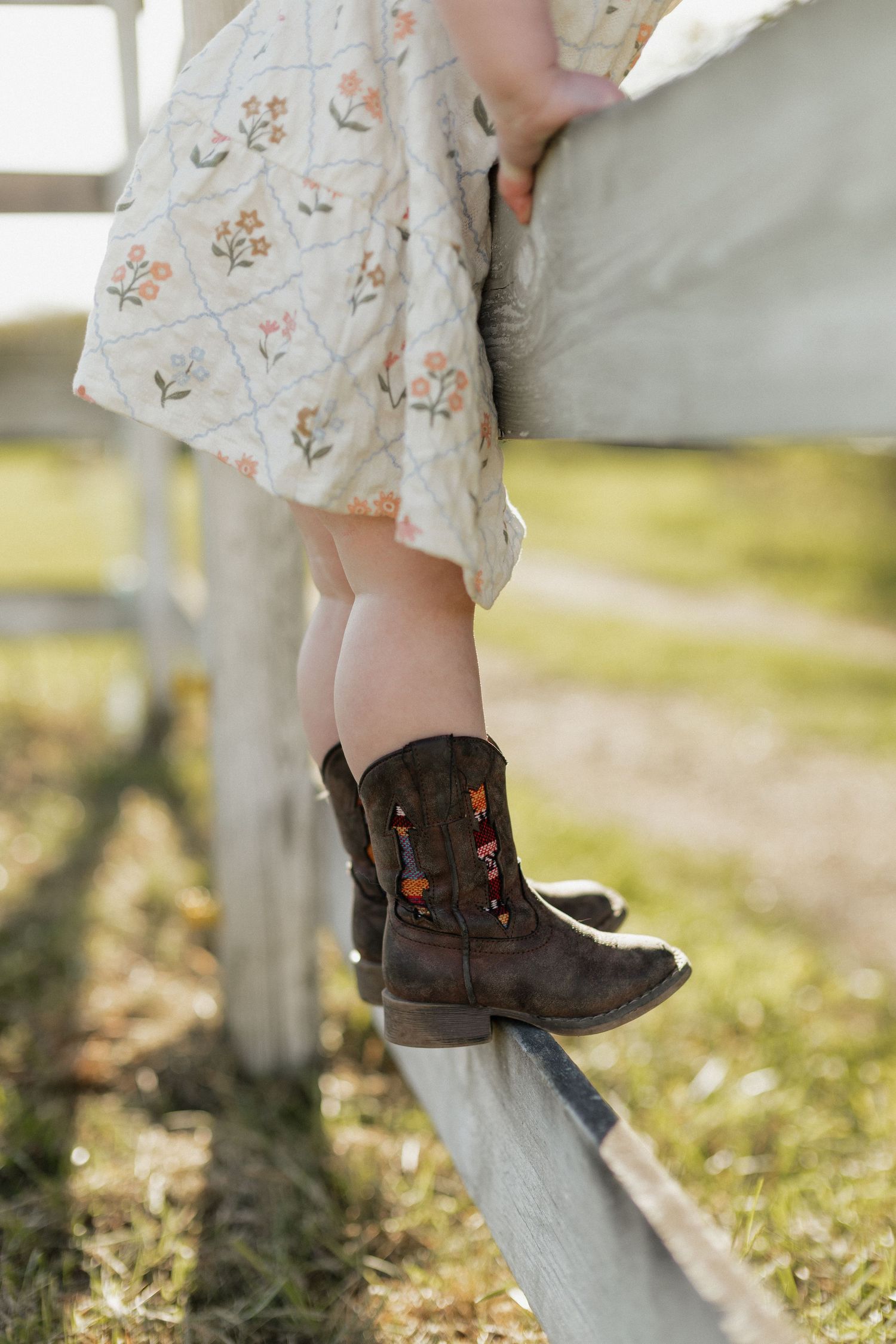 Child in floral dress and embroidered cowboy boots sitting on a white fence rail outdoors.