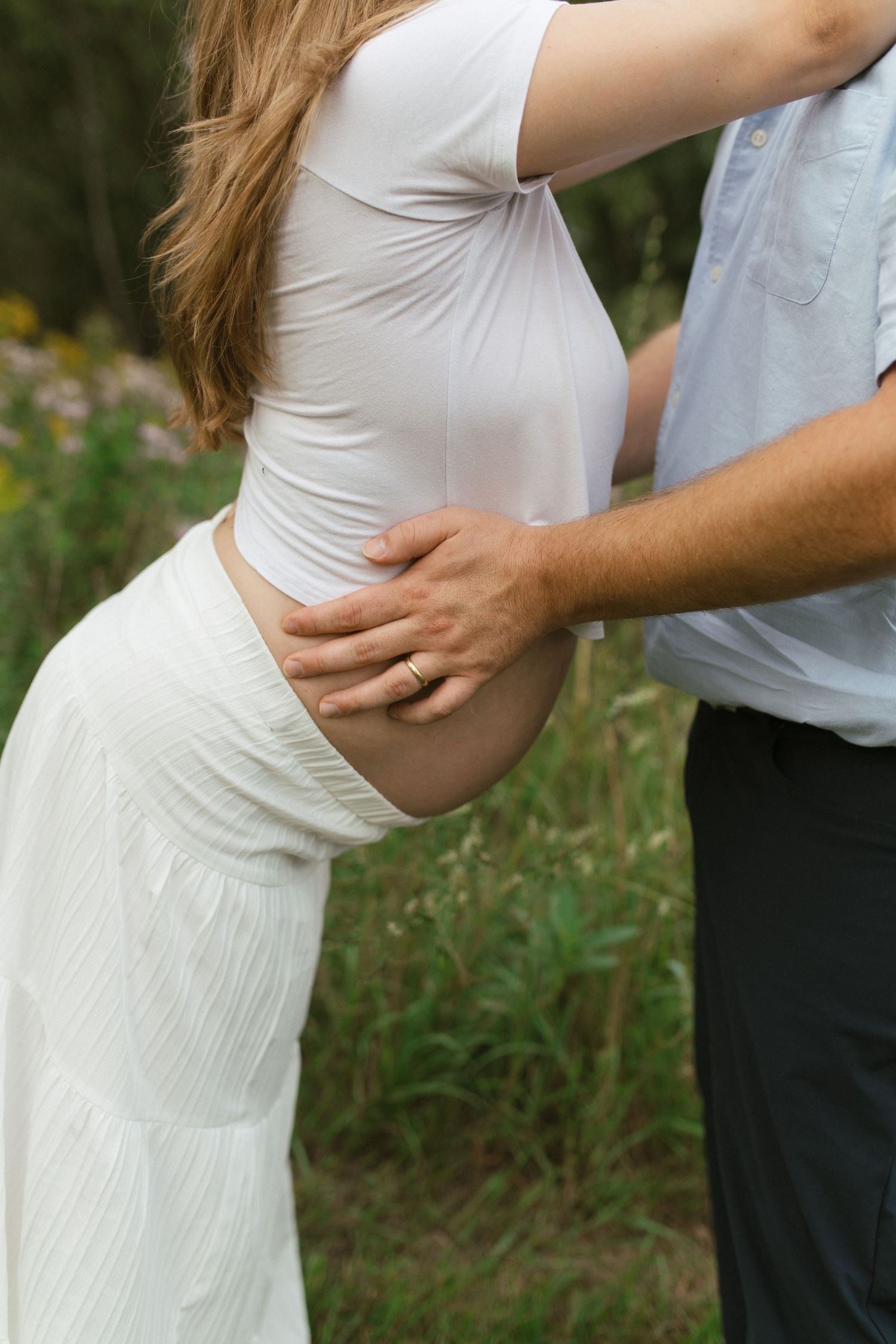 A man tenderly holds a pregnant woman's baby bump outdoors, both dressed in white near wildflowers.