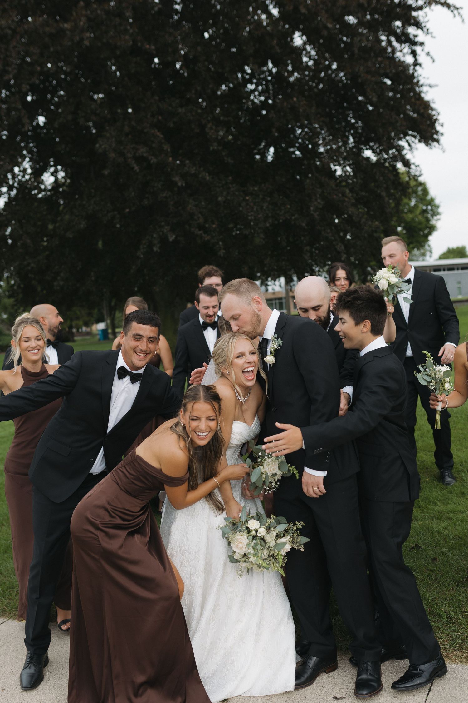 Wedding party laughing together outdoors, bride in white gown holding bouquet, bridesmaids in brown dresses, groomsmen in black tuxedos.