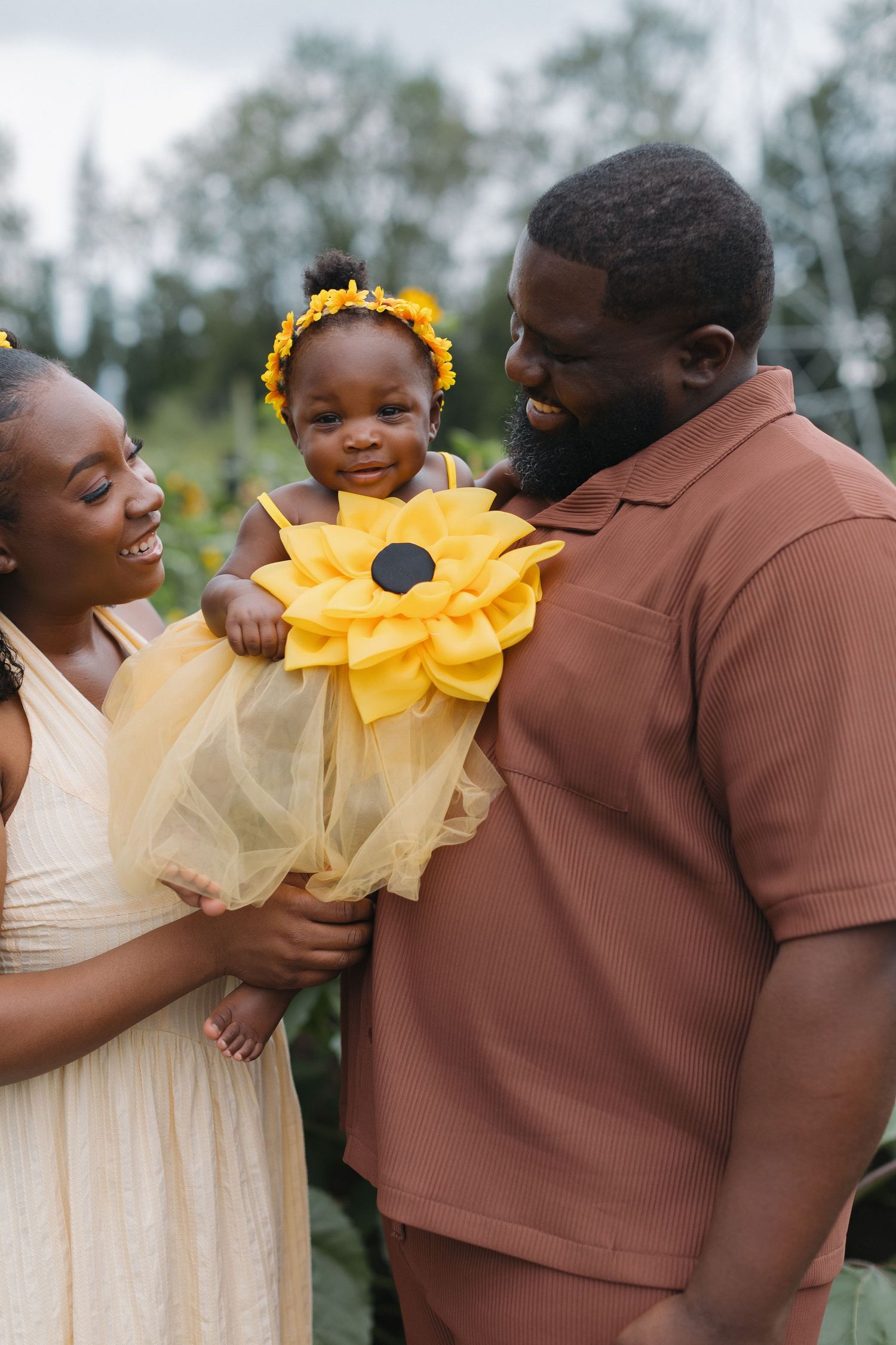 Happy Black family outdoors, baby in yellow sunflower costume with floral headband, parents smiling lovingly.
