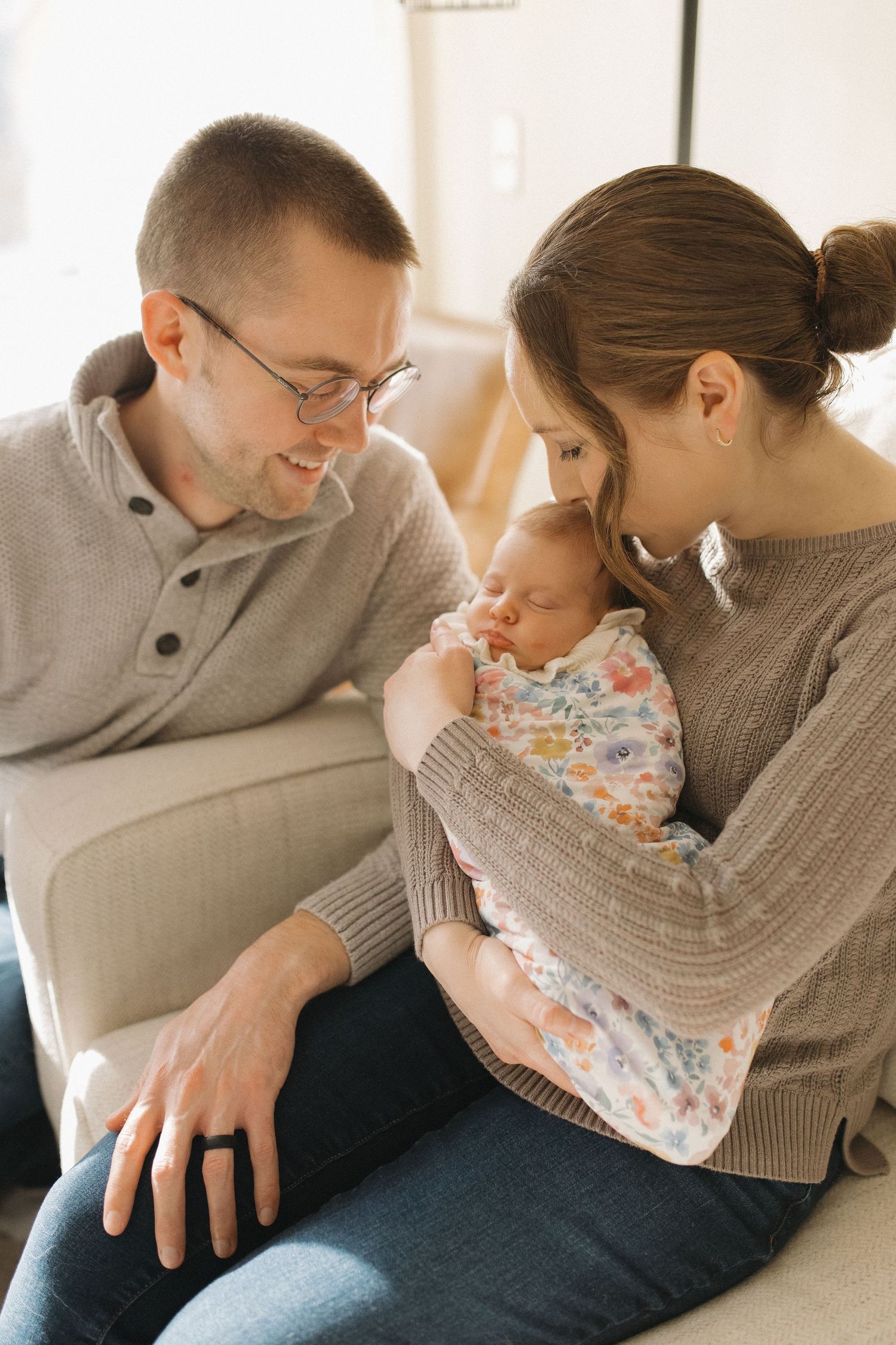 New parents sitting together, lovingly gazing at their sleeping newborn baby wrapped in a floral swaddle blanket.