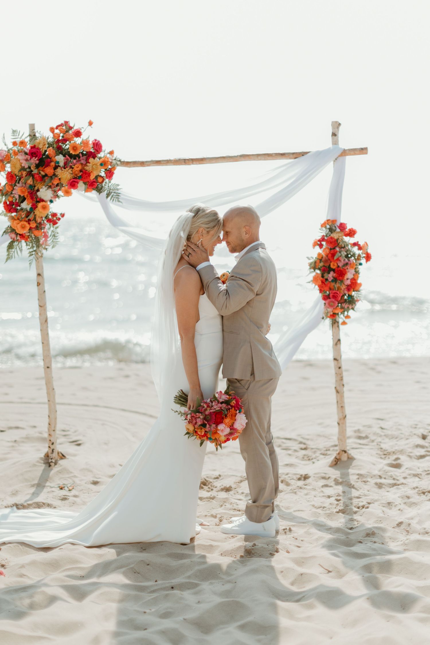 Beach wedding ceremony setup with coral floral arrangements and white fabric arch.