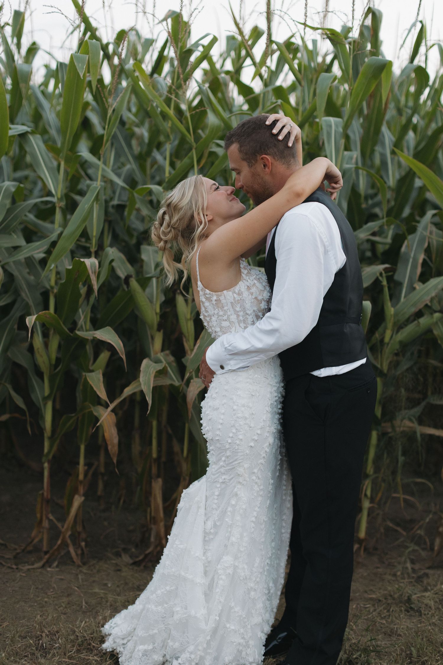 Bride and groom embracing and kissing in front of a lush green cornfield in rustic outdoor wedding photo.