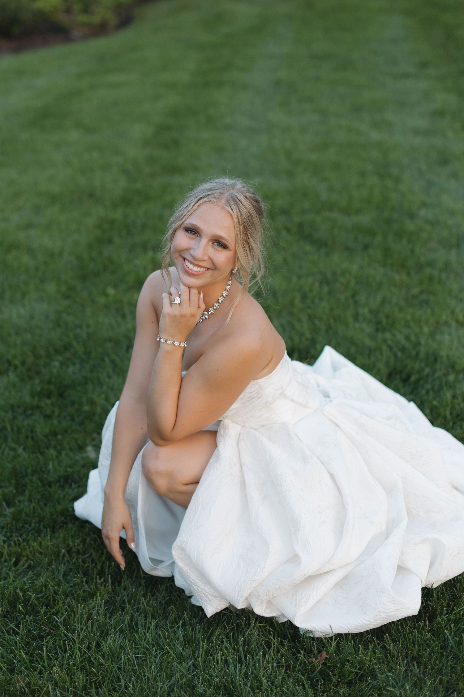Smiling blonde bride in white strapless gown sitting on green grass, wearing pearl jewelry and engagement ring.