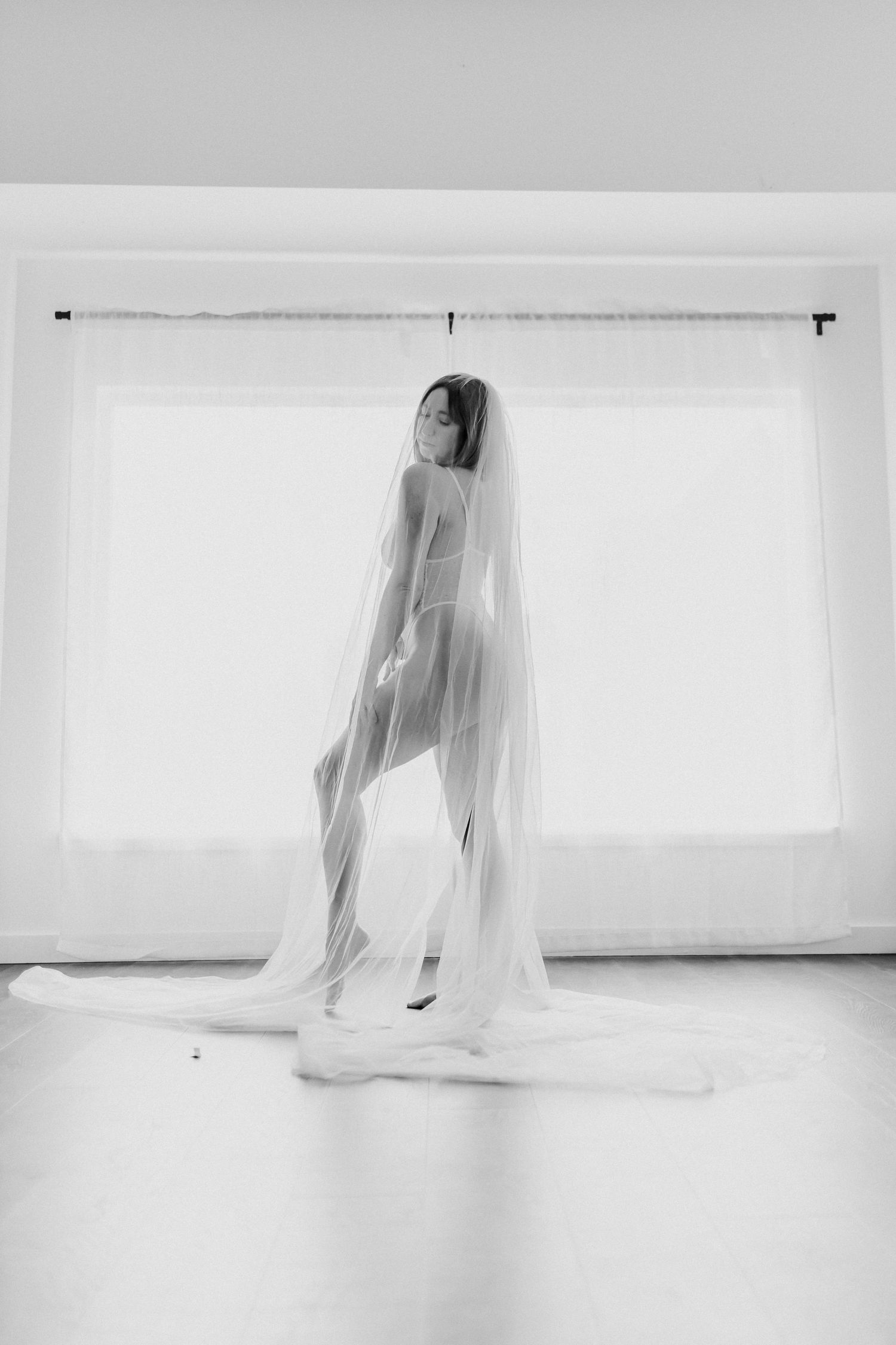 A minimalist black and white boudoir photo of a bride wrapped in a flowing veil in an empty white room.