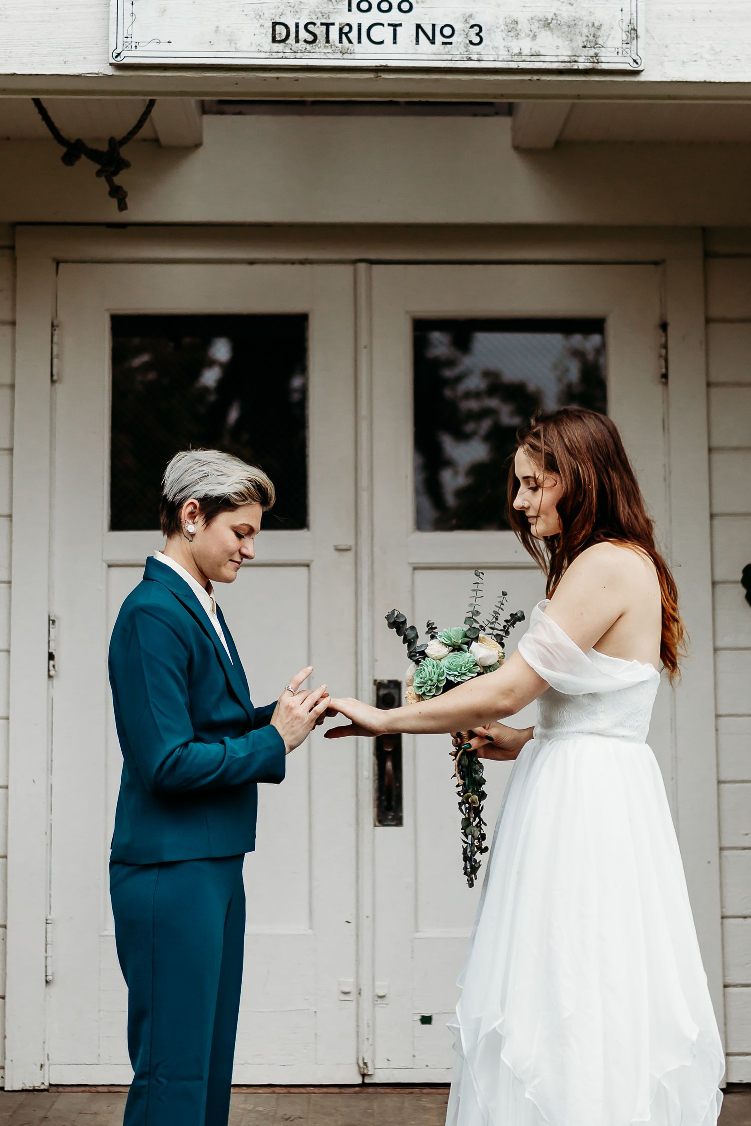 Couple exchanging rings outside historic white schoolhouse building.