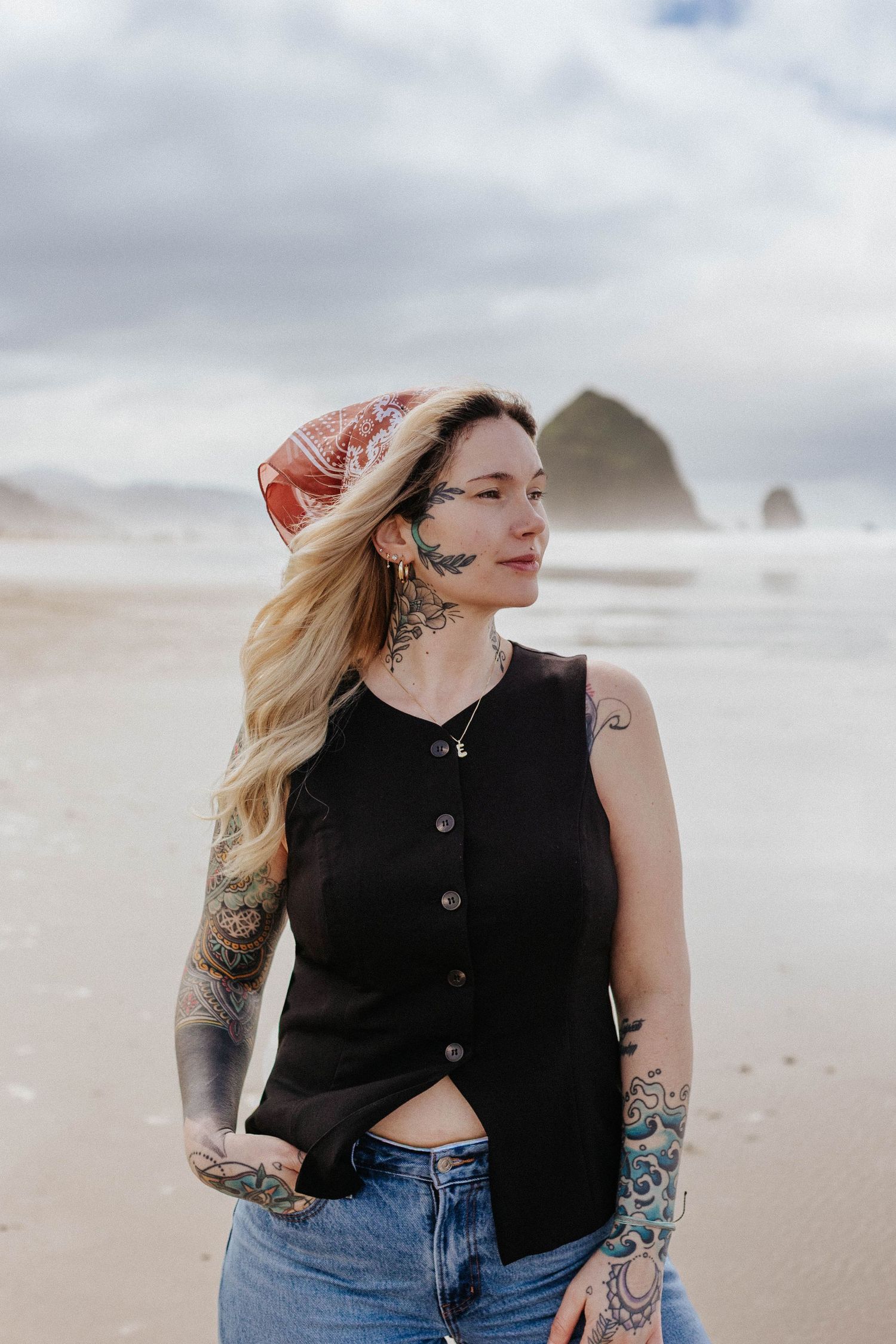 Tattooed blonde woman in black vest and red bandana gazes sideways on misty beach with rock formation.