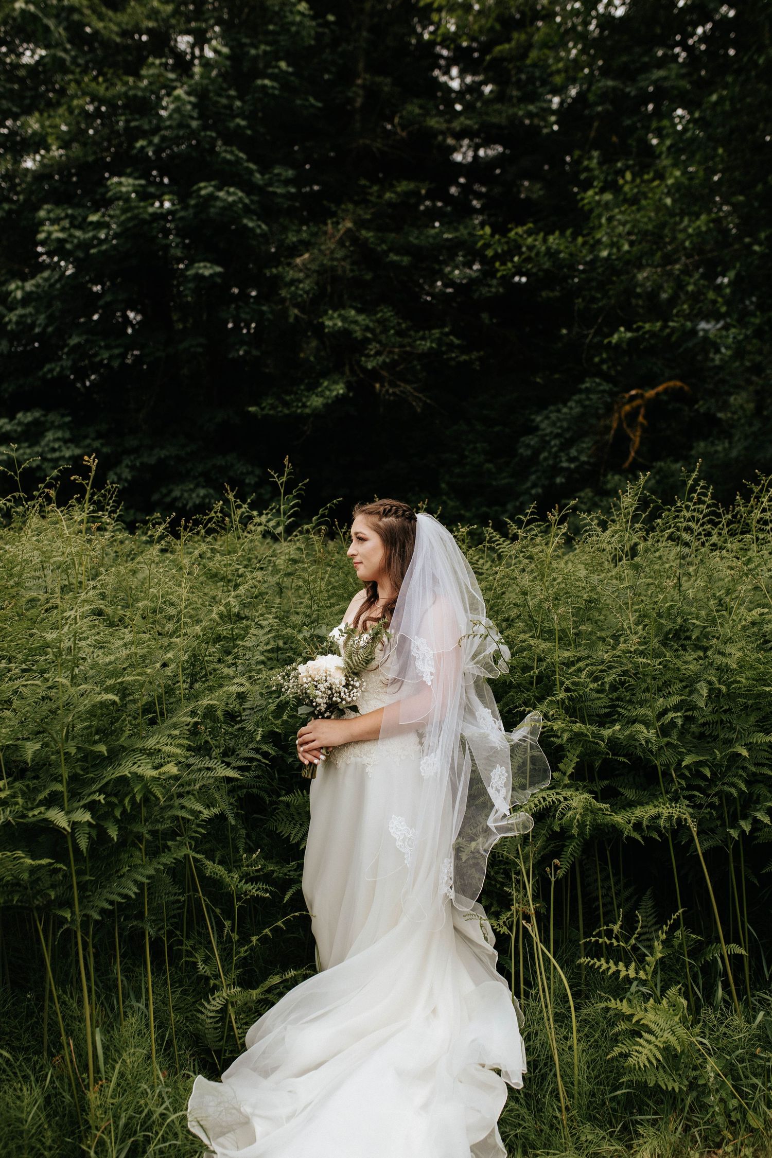 Bride in flowing white gown and veil holds a bouquet while standing among lush green ferns outdoors.