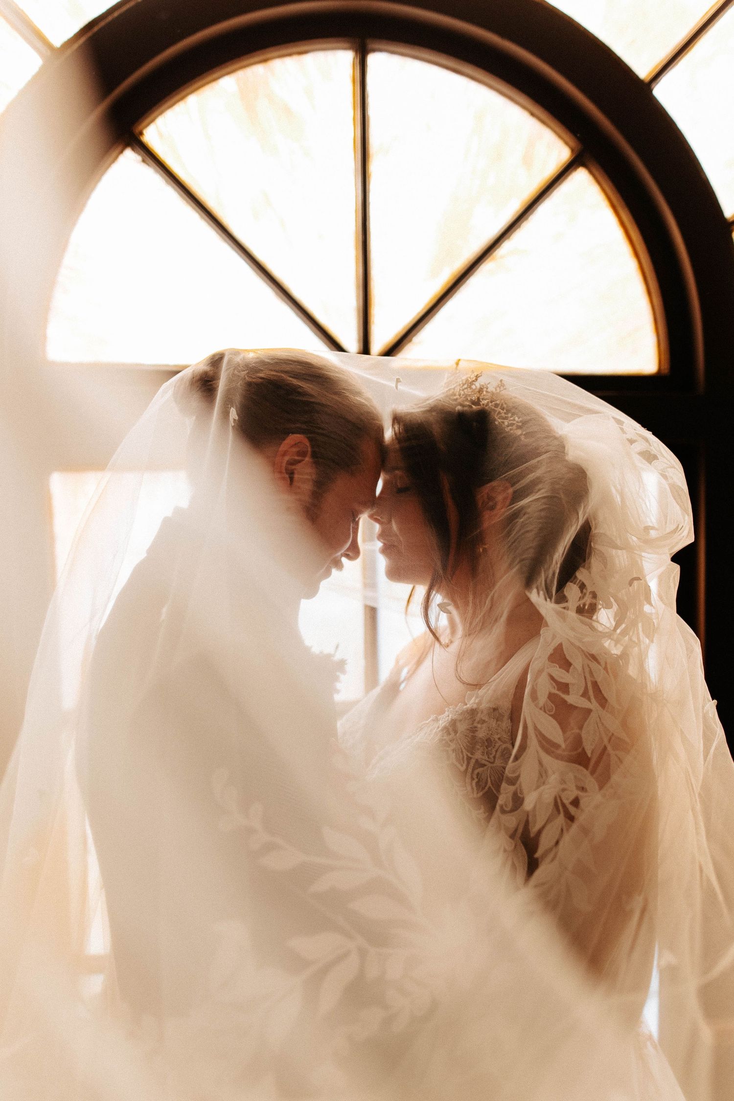 Romantic couple leans together under flowing veil before arched window in soft golden backlit light.