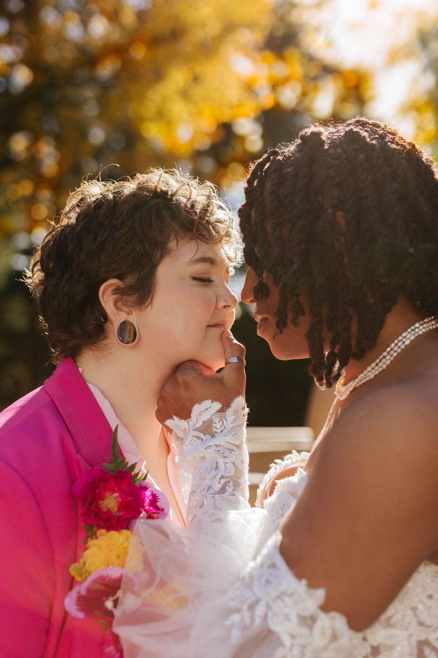 Two brides share a tender moment outdoors in autumn, one in a pink blazer and one in a white lace gown.