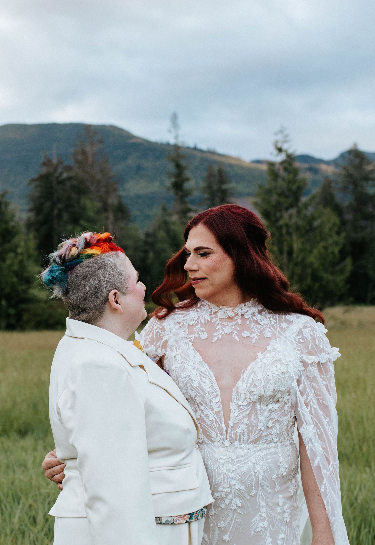 Two newlyweds sharing an intimate moment in a meadow with mountains and evergreen trees in the background.