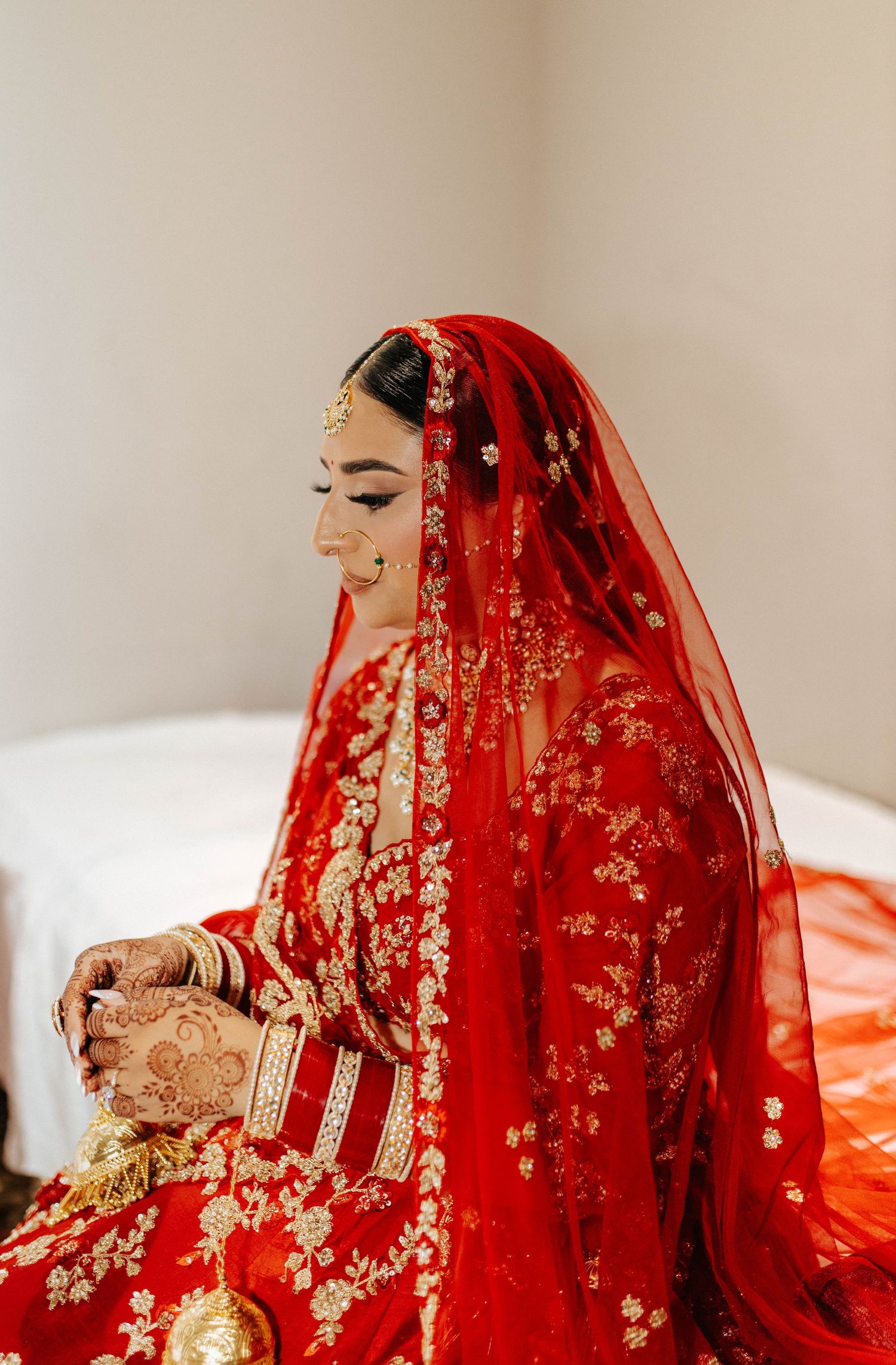 A bride in a traditional red Indian wedding dress with intricate gold embroidery and henna-decorated hands sits in contemplation.
