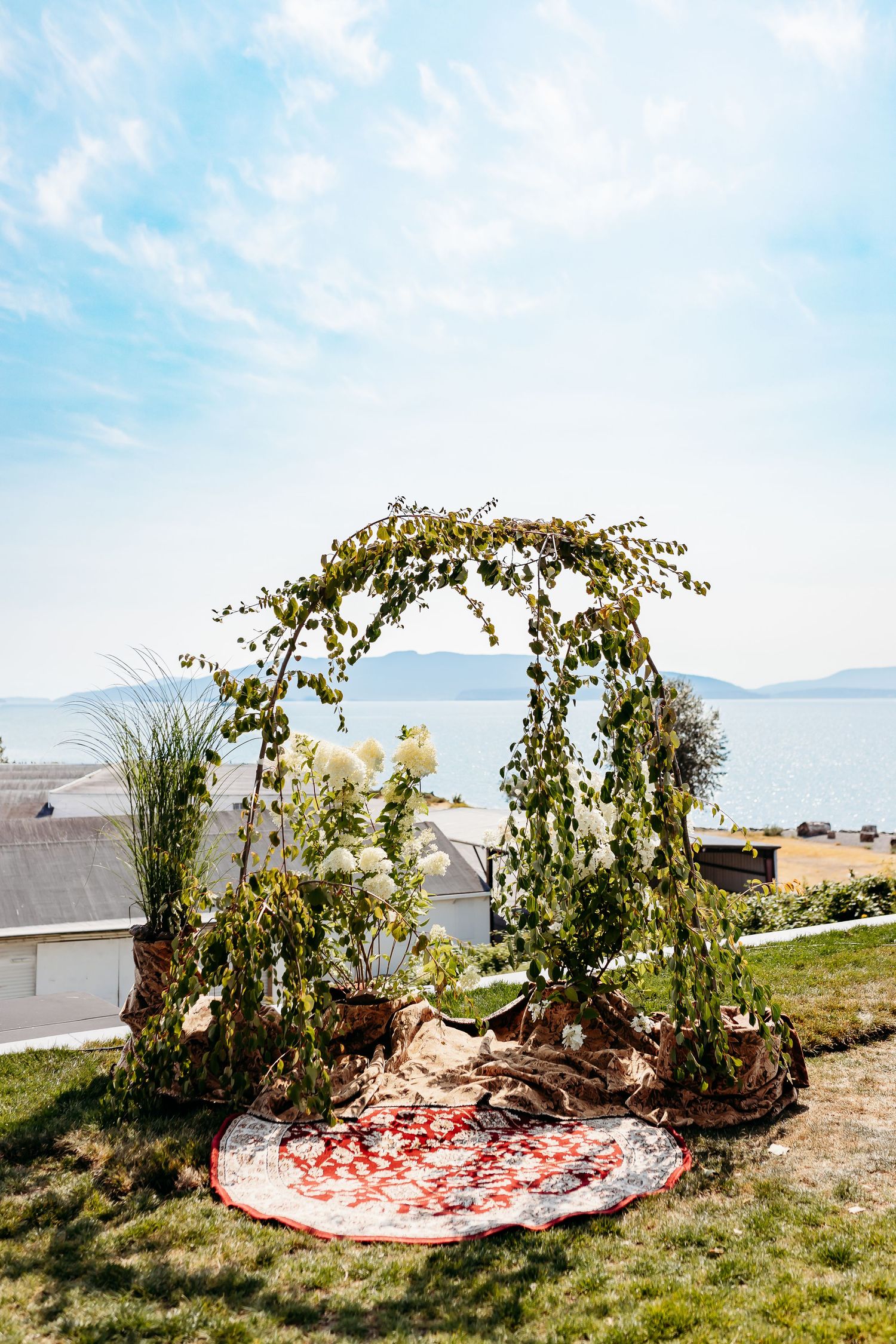 Wedding arch decorated with greenery and flowers overlooking a scenic ocean view with rose petals scattered below.