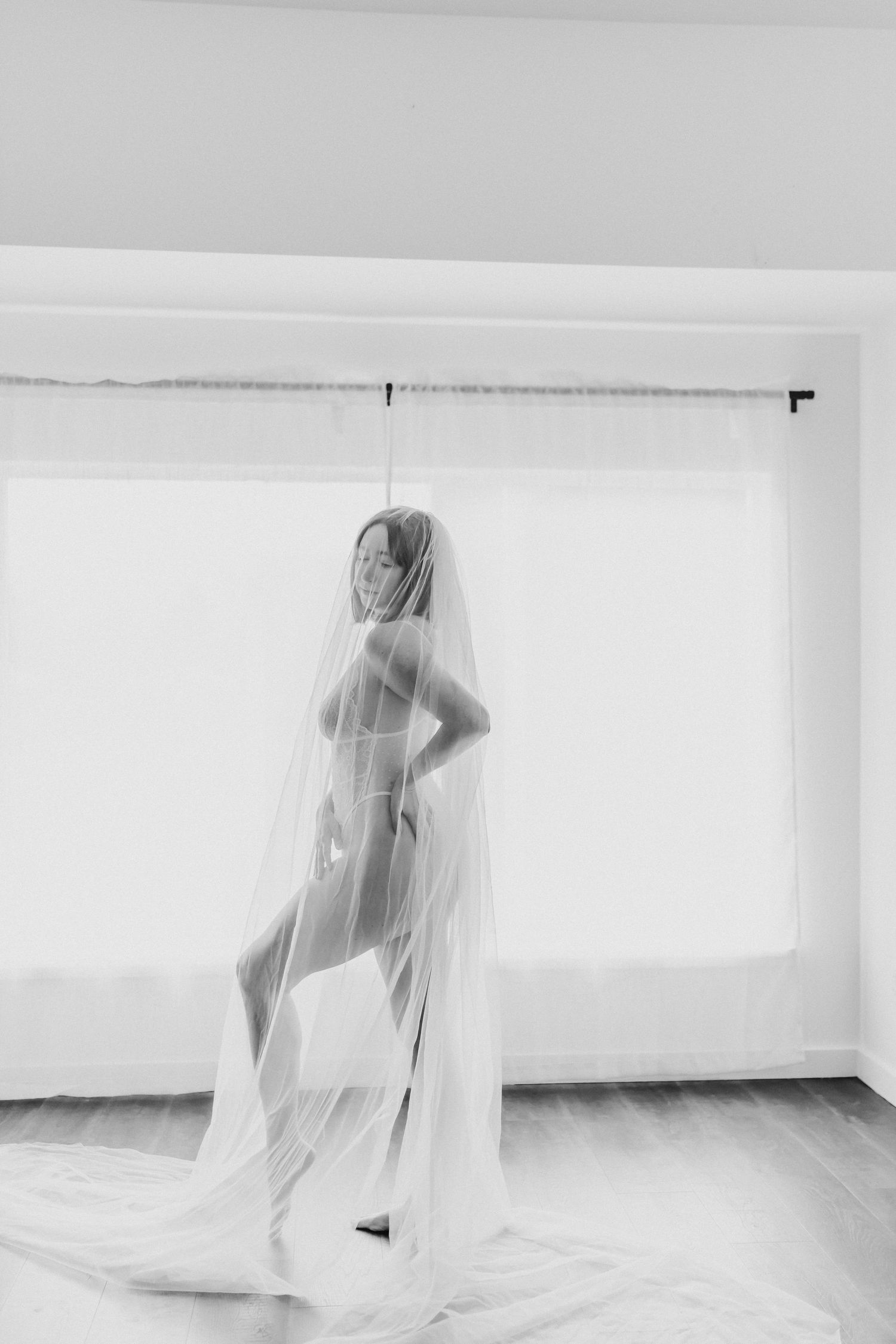 Artistic black and white boudoir photo with flowing sheer veil and feminine silhouette against bright window light.