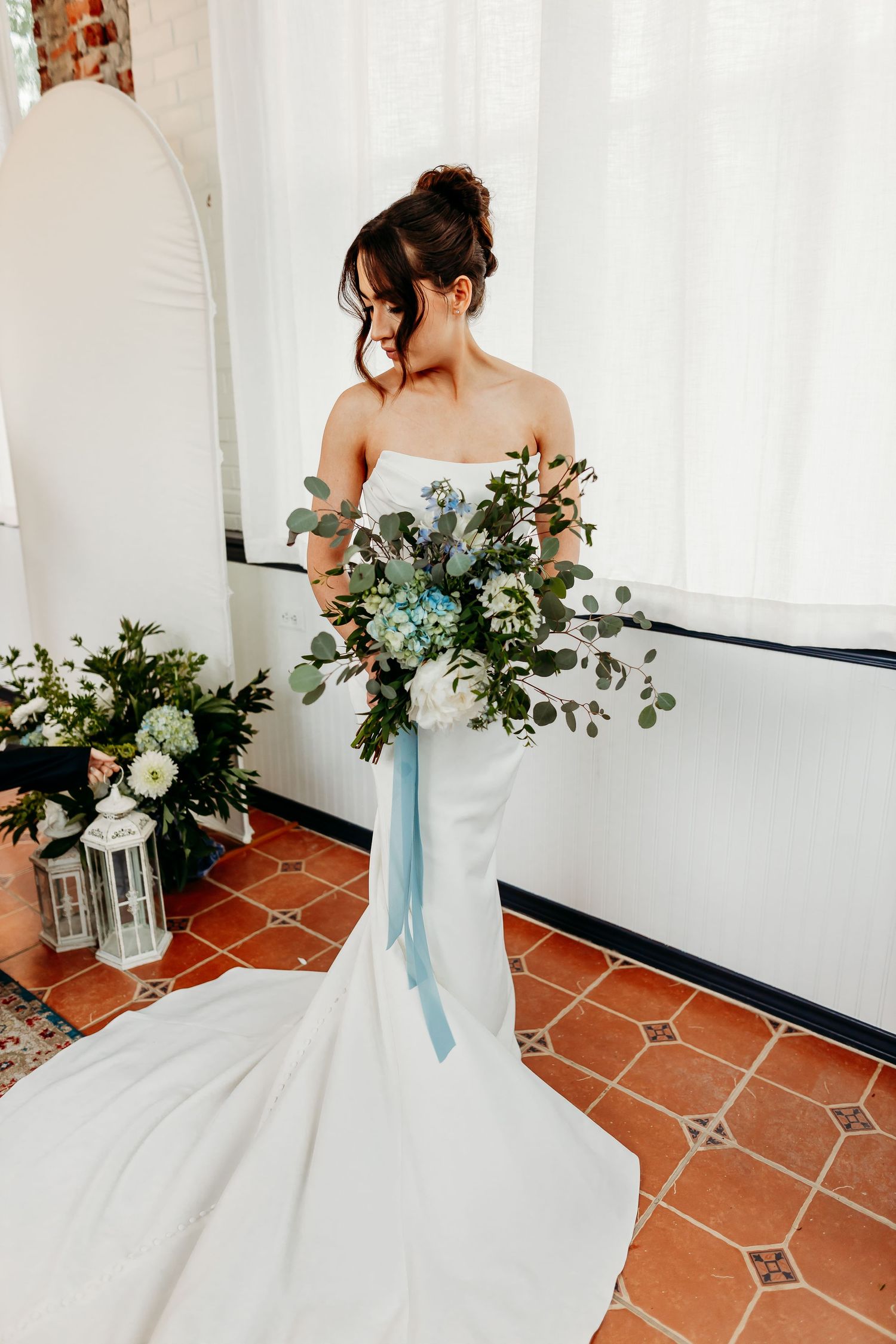A bride in a fitted white gown holding a blue and white bouquet with trailing ribbons.