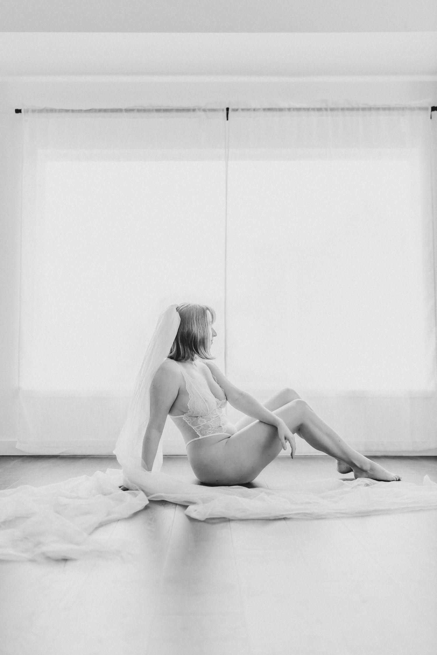 Minimalist black and white boudoir photograph with delicate veil and romantic pose on hardwood floor.