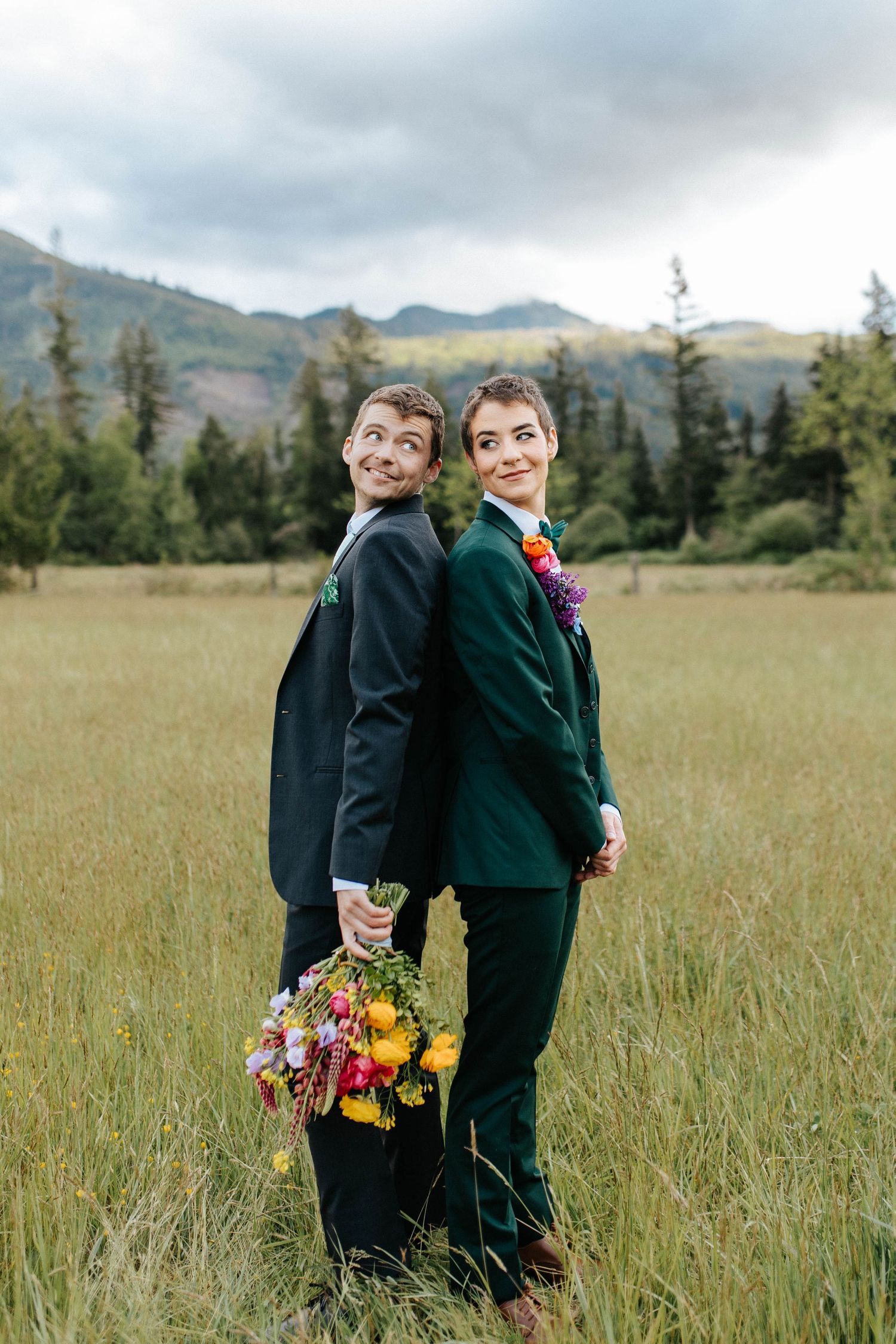 Two people in formal attire standing back-to-back holding colorful bouquet in mountain meadow with forest backdrop.