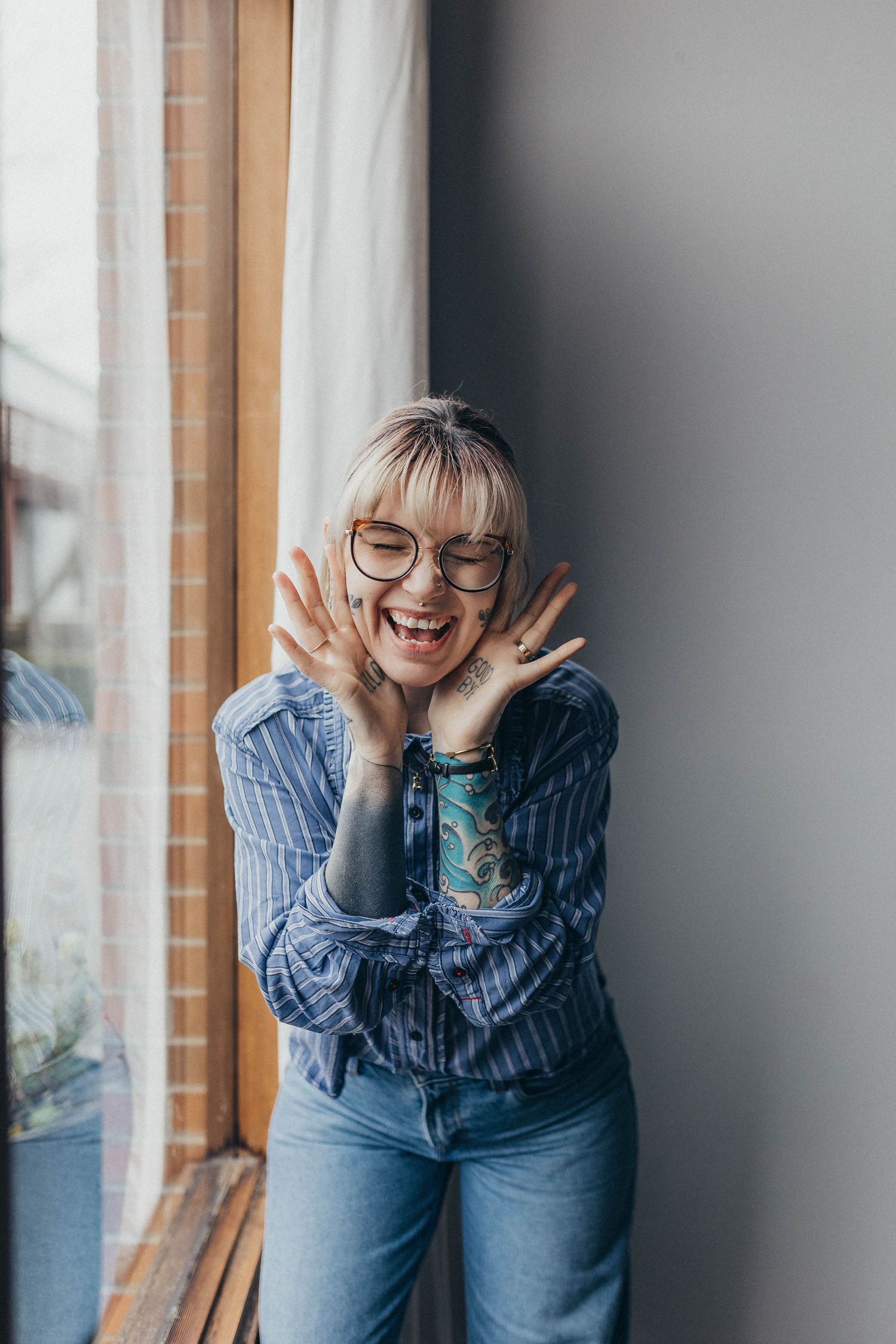Tattooed blonde woman with glasses laughs joyfully near a window in a blue striped shirt.