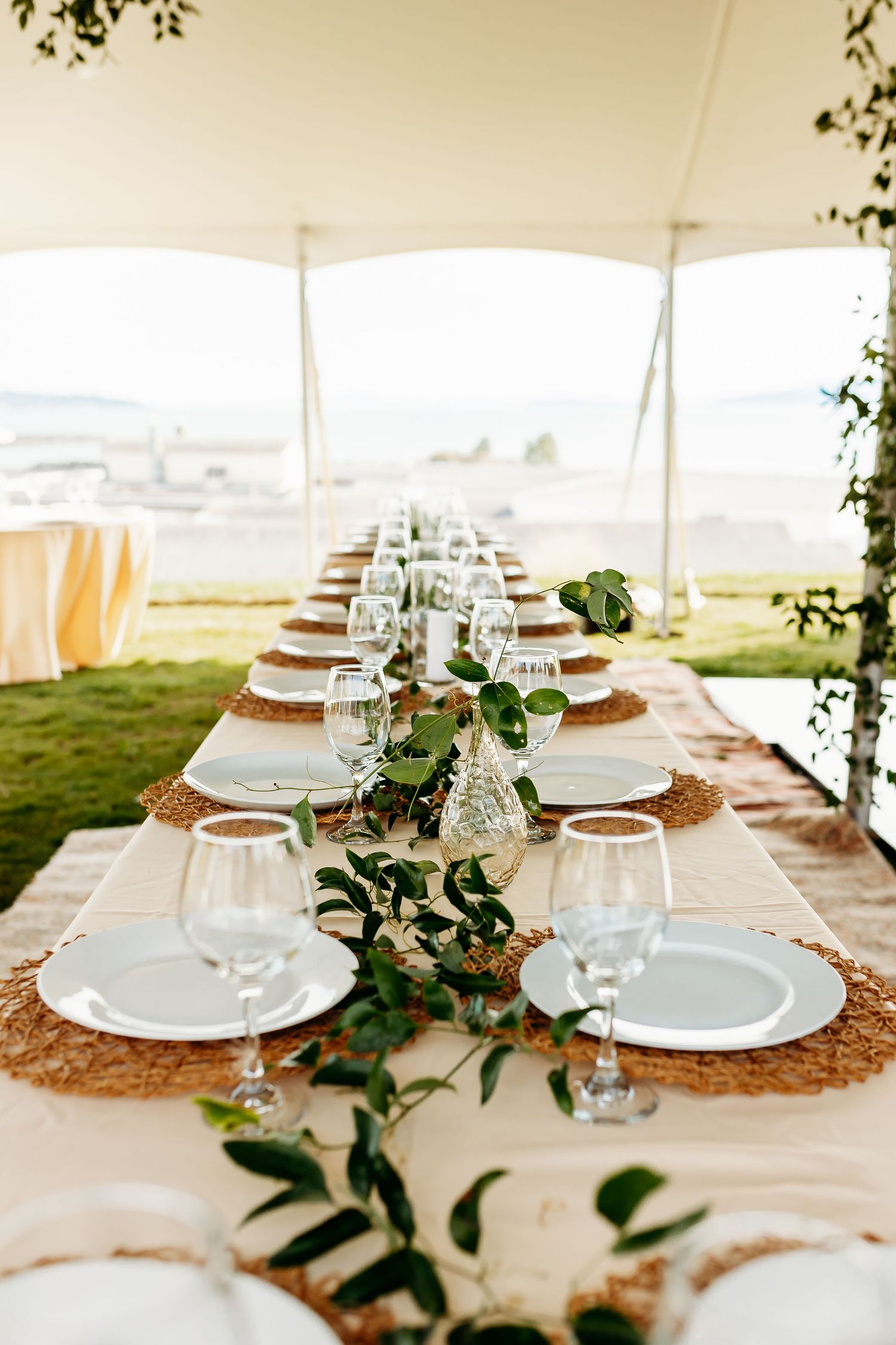 Elegant outdoor wedding table setting with eucalyptus garland, white plates and cork placemats under a white tent canopy.