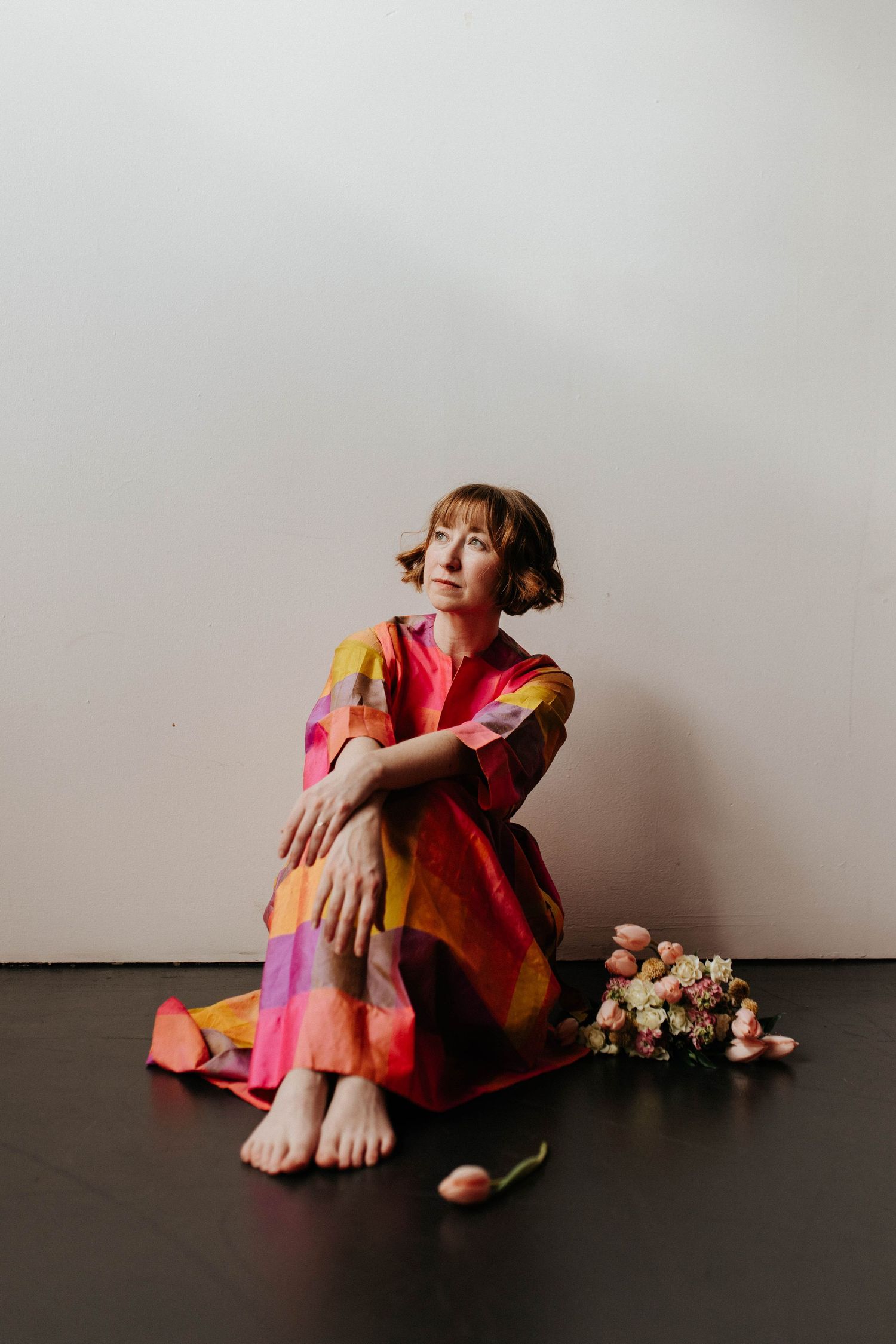 A figure in a vibrant floral dress sits barefoot on a dark floor with flowers nearby against a white background.