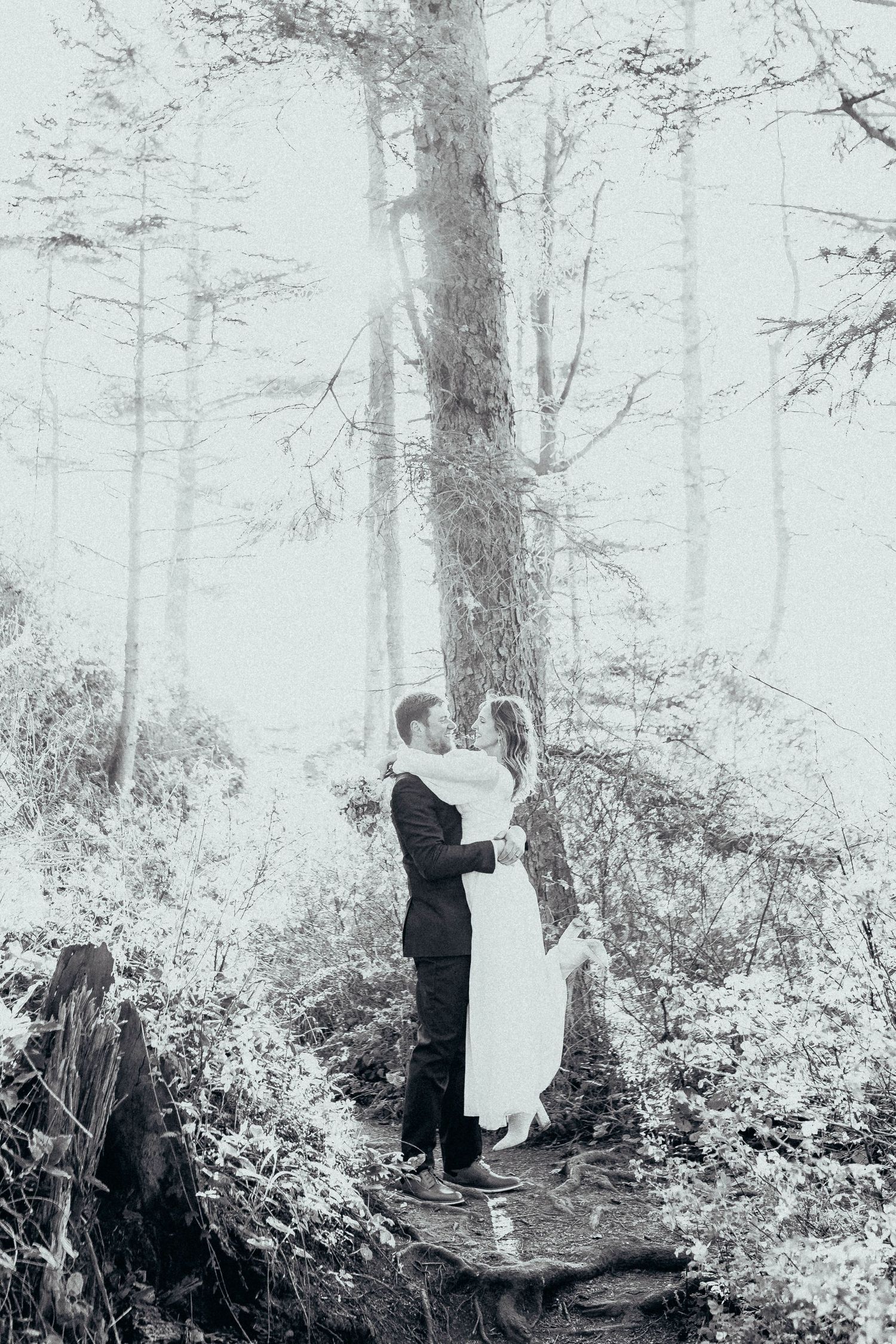 Black and white photo of couple embracing and kissing among tall trees in a misty forest setting.