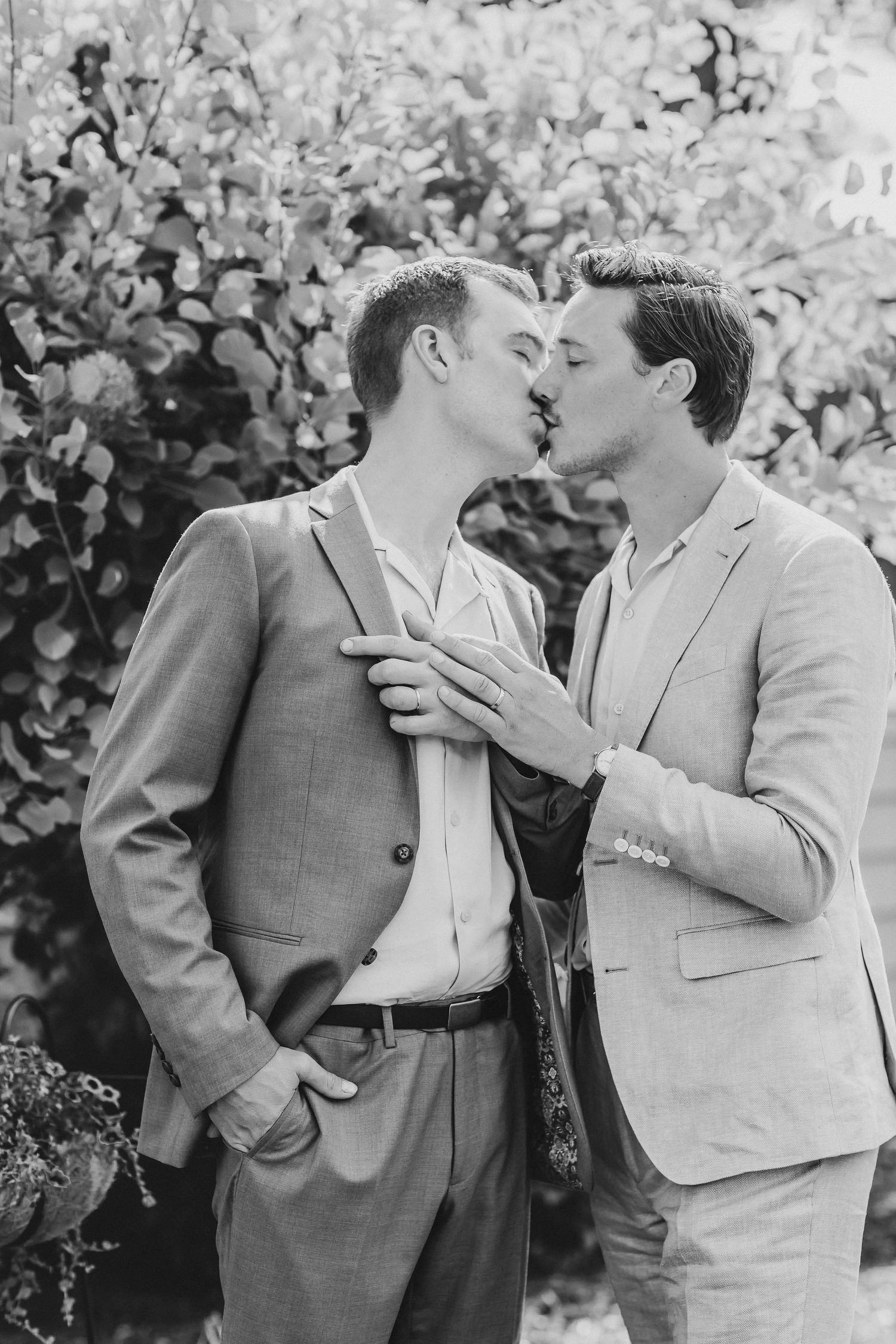 Two people in suits share a tender romantic moment while standing in front of blooming foliage in this black and white photo.