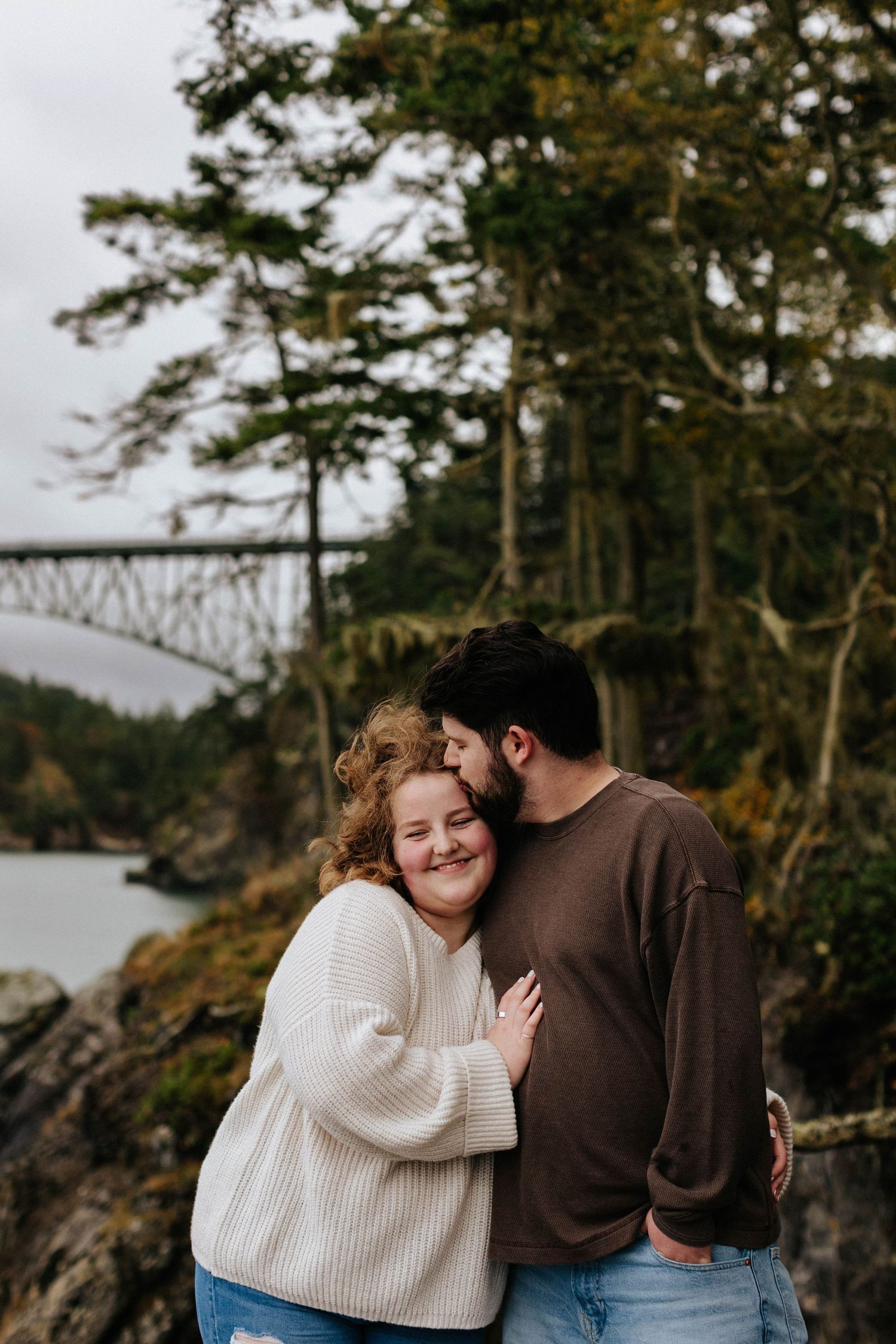 Couple embracing outdoors near Deception Pass bridge, man kissing woman's cheek as she smiles warmly.