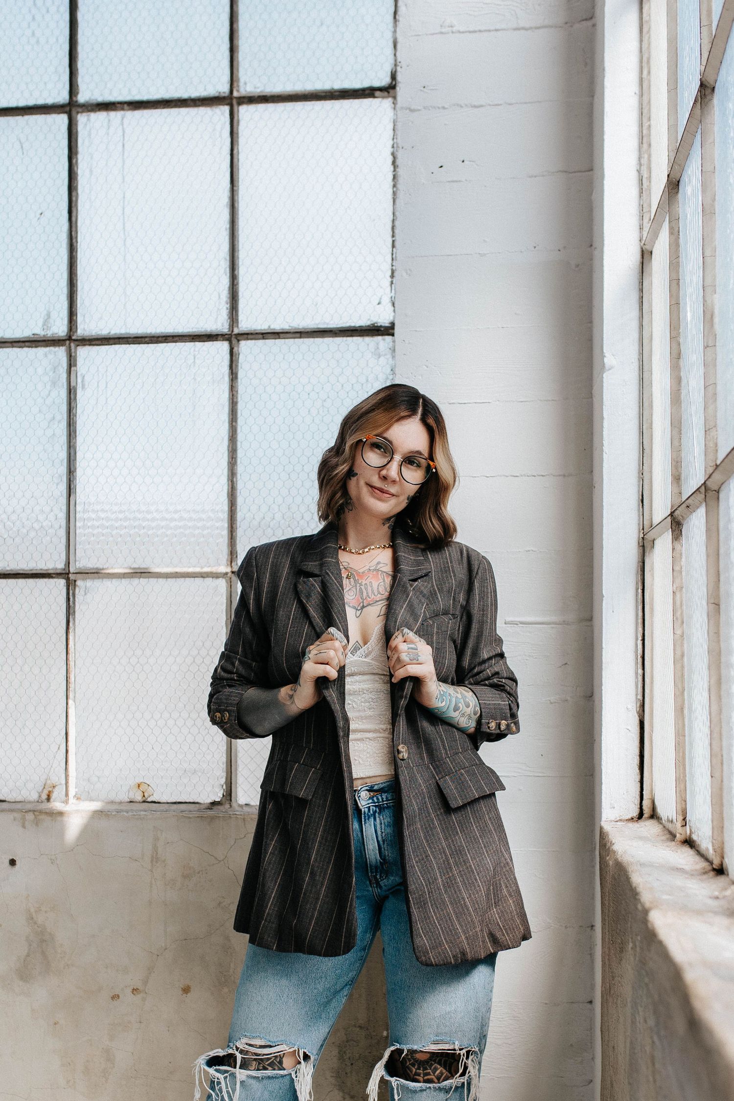 Woman in striped blazer, lace top, and ripped jeans poses by industrial windows with natural light.