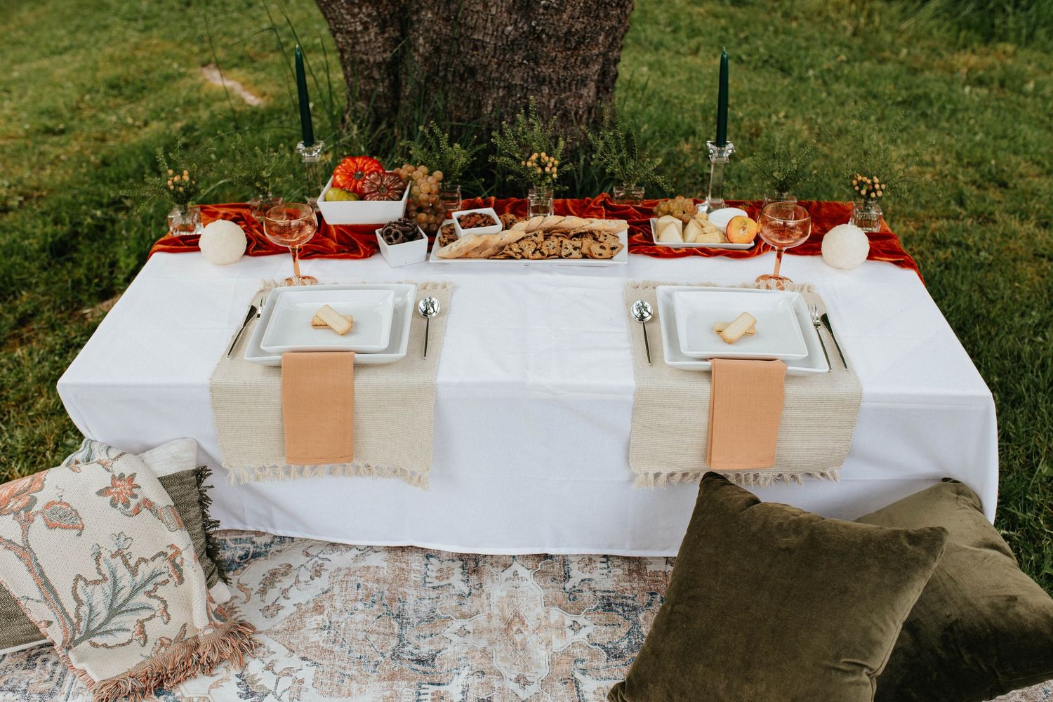 Romantic outdoor picnic setup with white tablecloth, peach accents, and cushions under a tree with red floral decorations.