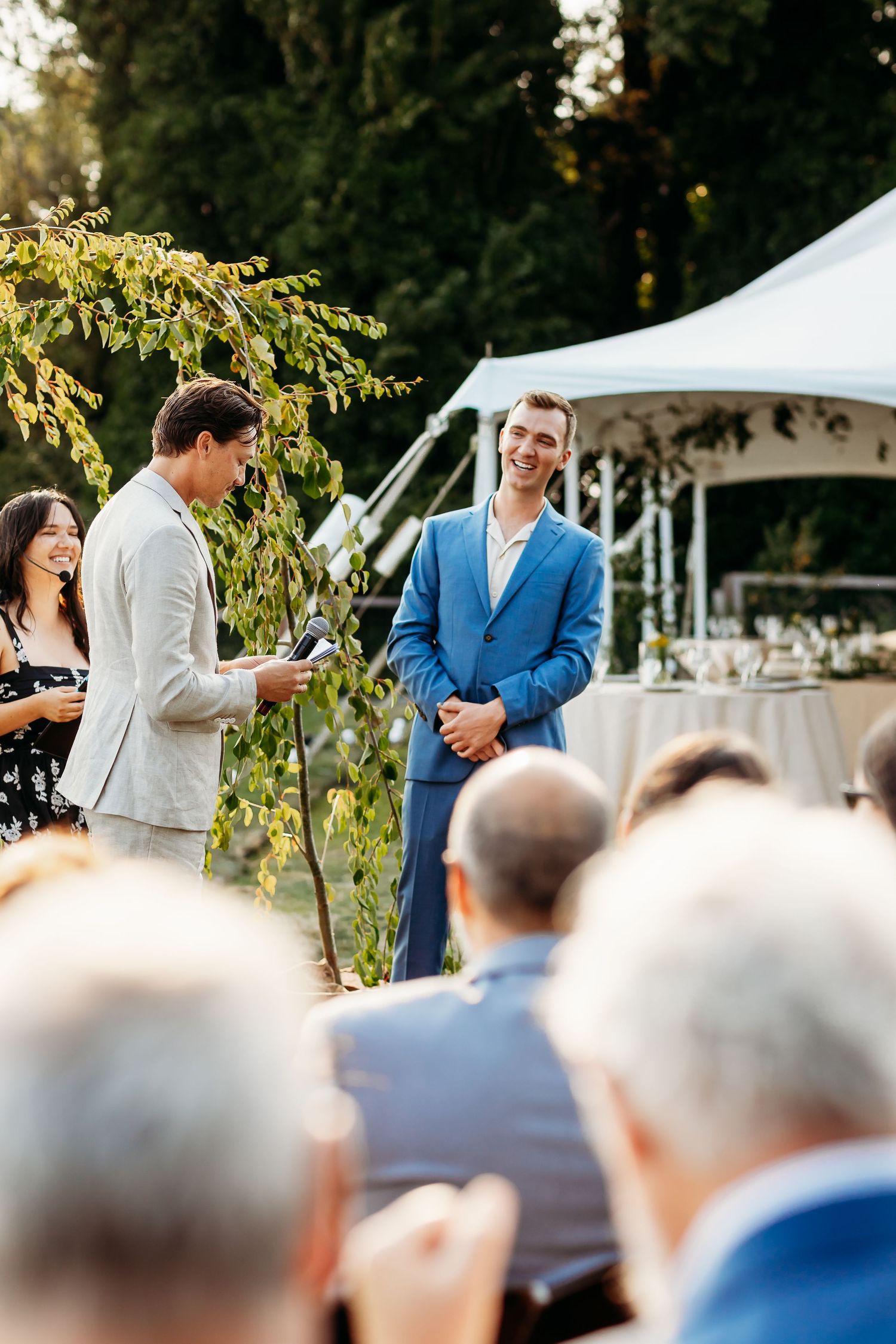 An outdoor wedding ceremony takes place under a white tent with guests seated and greenery in the background.