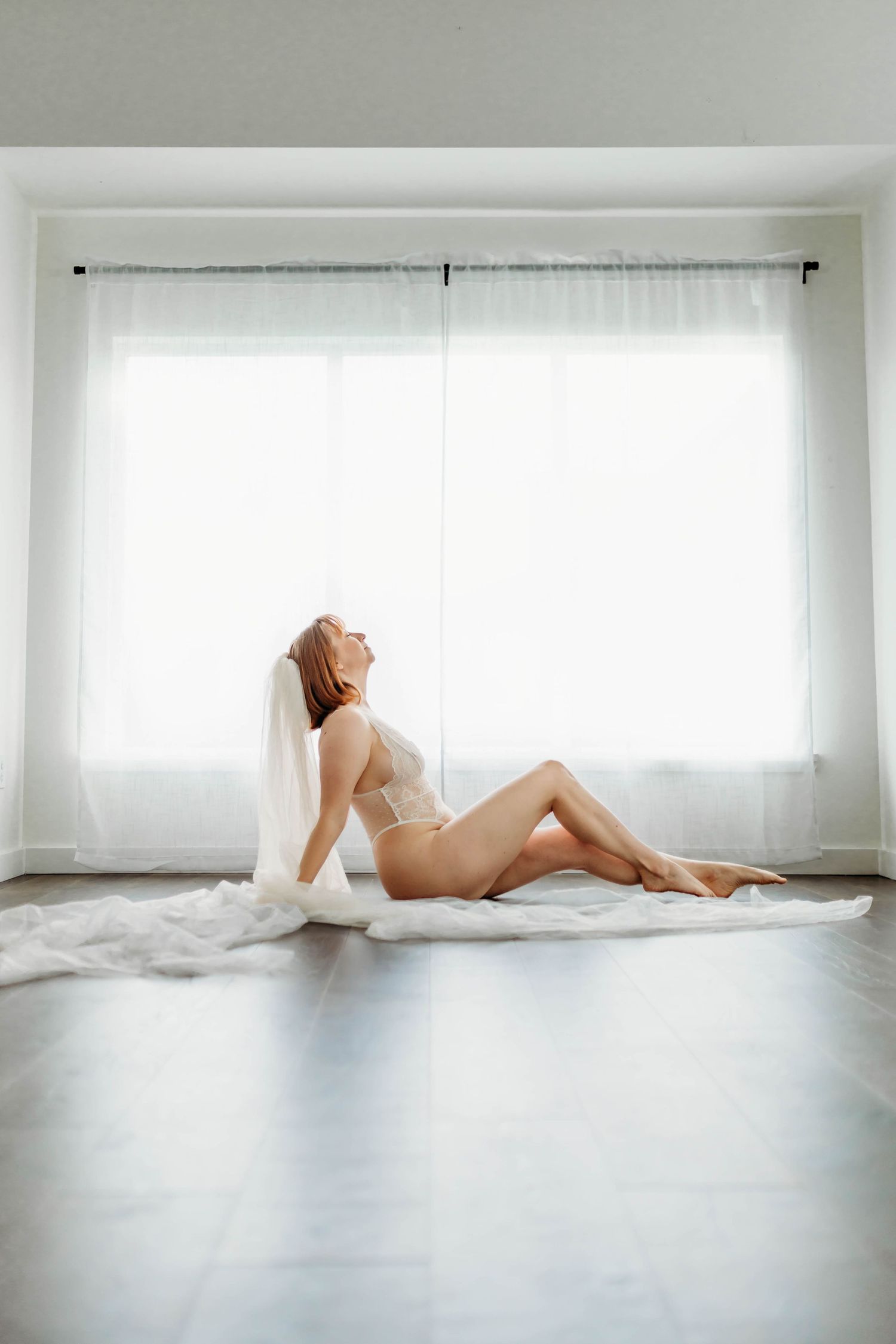 Figure in white bridal veil reclines gracefully against bright window in minimalist boudoir setting.