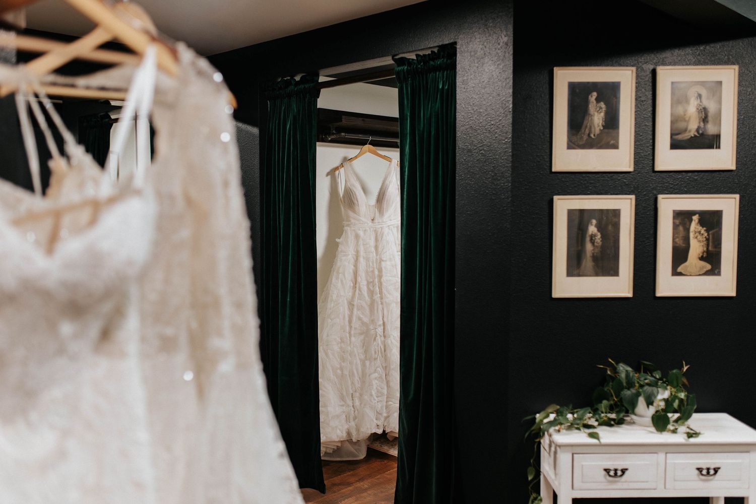 Two wedding dresses hang elegantly in a dark-walled bridal suite with framed photos on the wall.