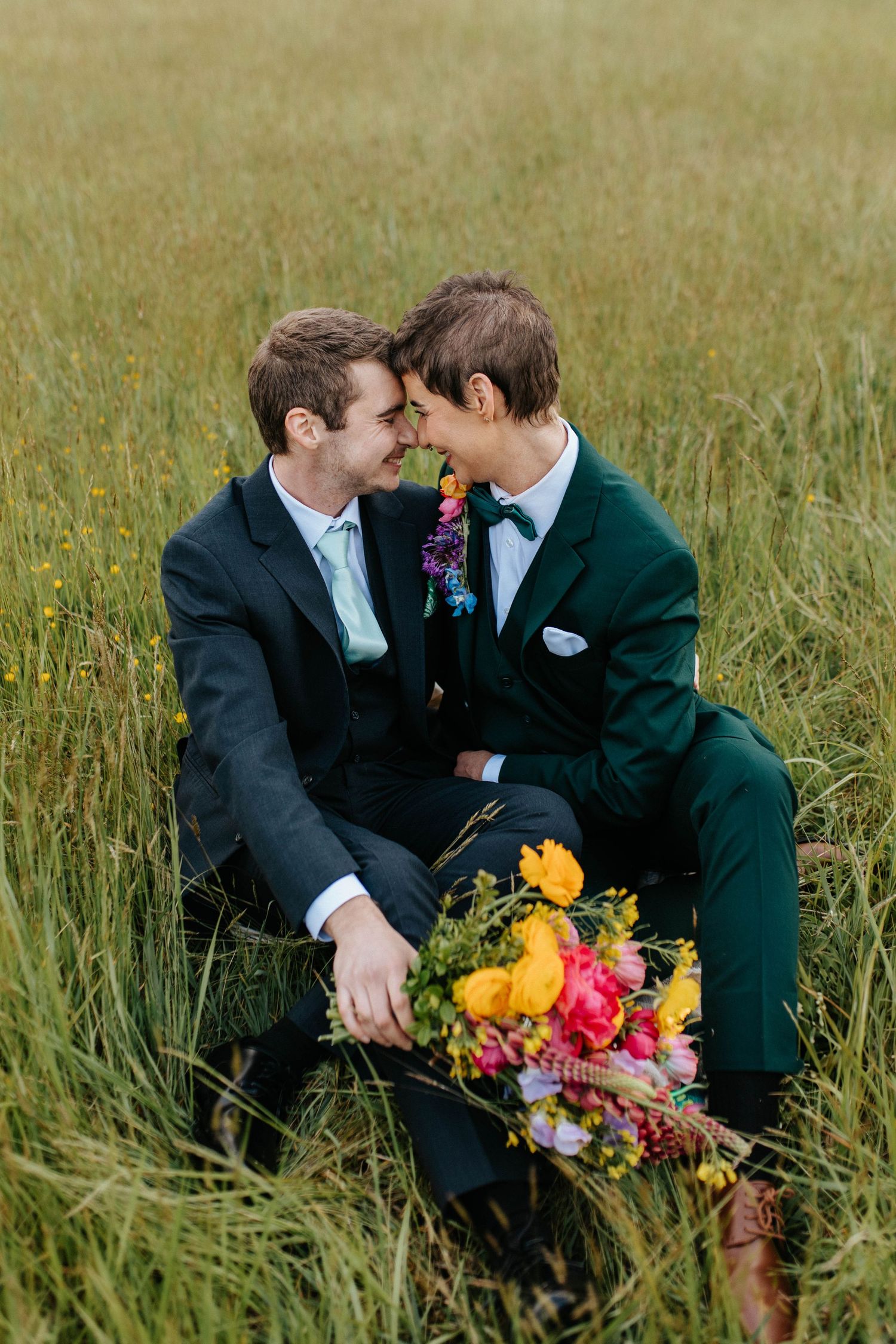 Two people in formal wear share an intimate moment while sitting in tall grass with a vibrant floral bouquet.