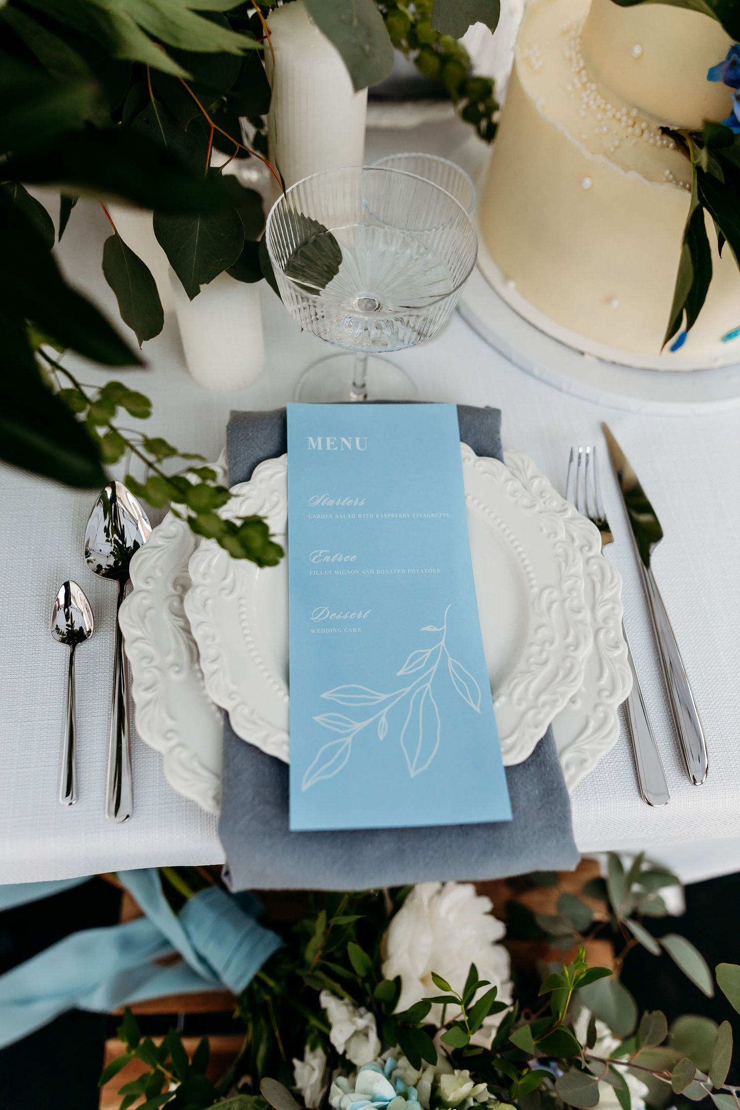 Elegant place setting featuring light blue menu card on gray napkin with white plate and floral decor.