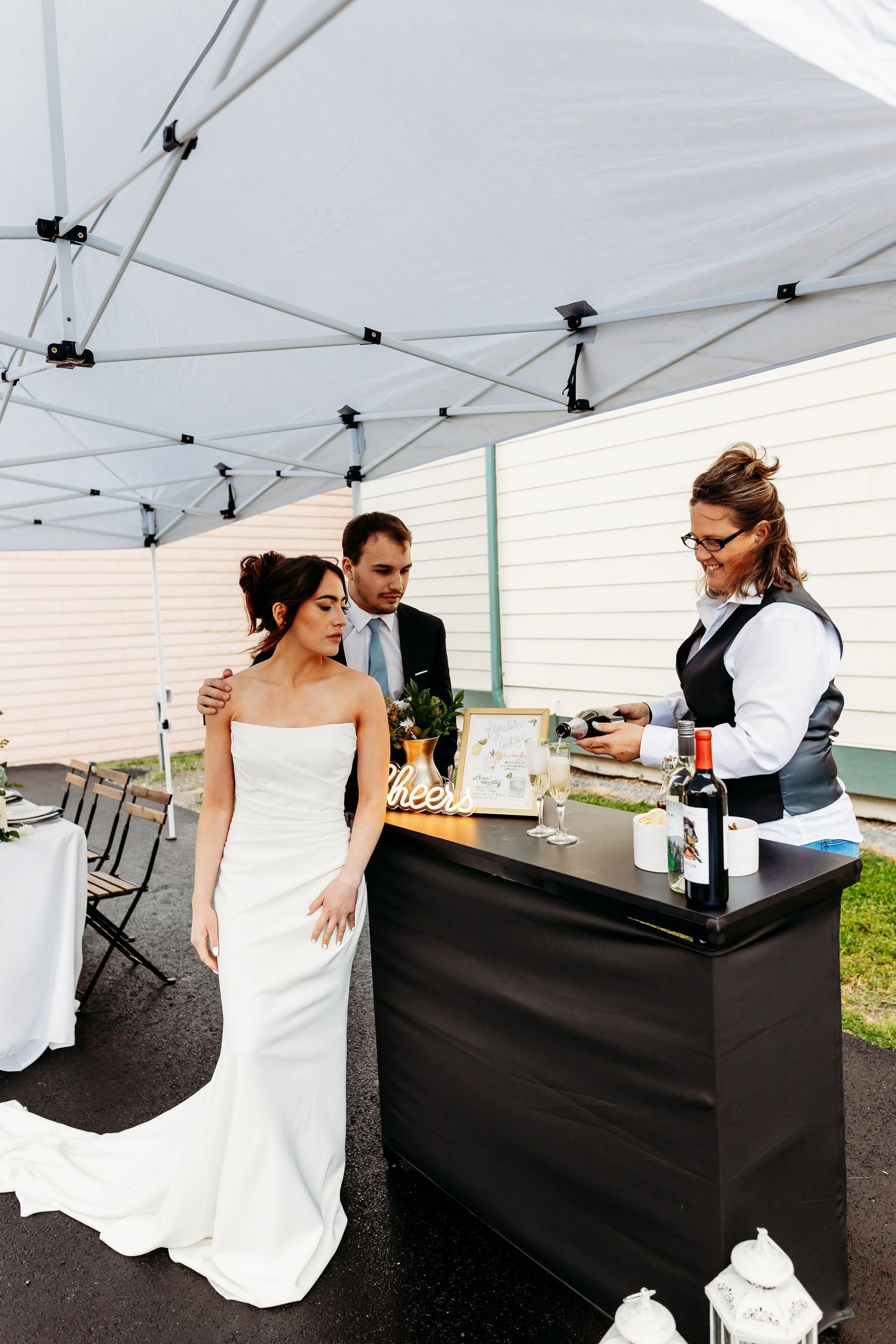 A couple in wedding attire stands at a portable bar under a white tent while a bartender serves drinks.
