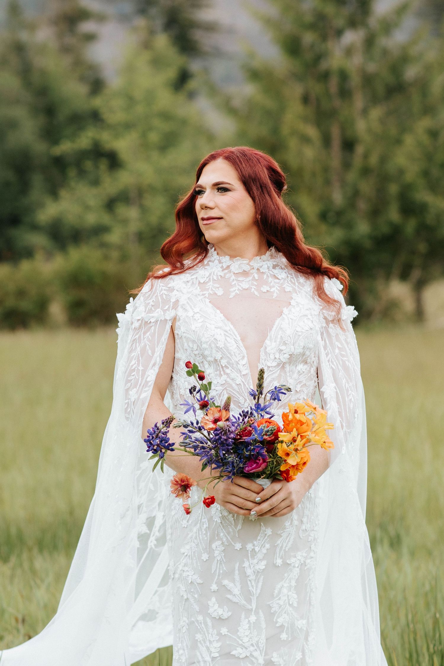 Bride in lace wedding dress holding colorful bouquet stands in mountain meadow surrounded by evergreen trees.