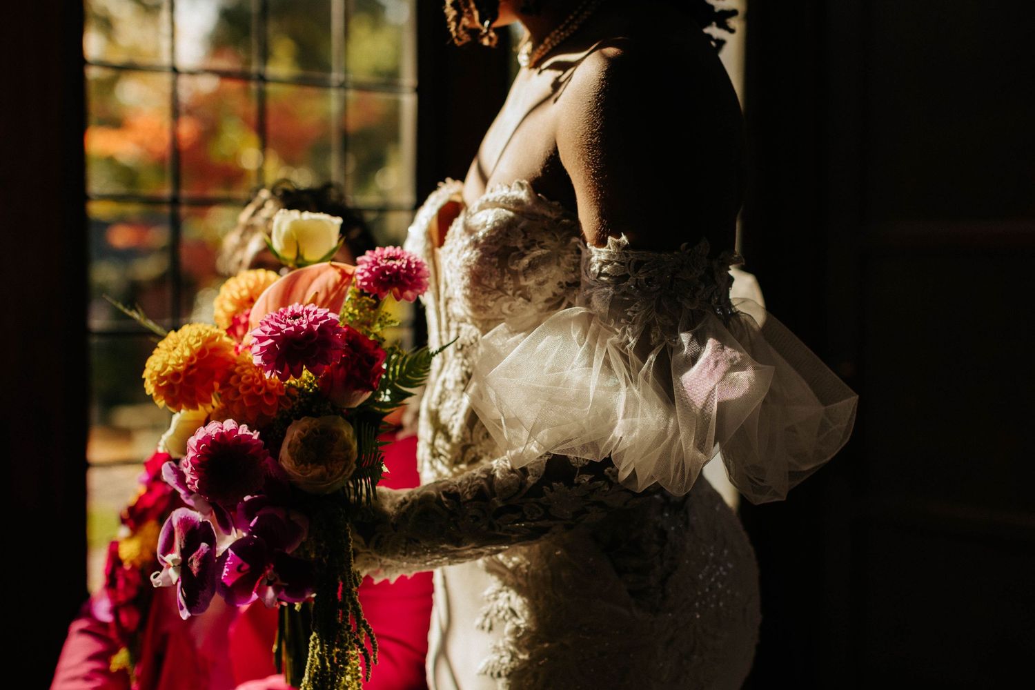 Bride in lace gown holding vibrant pink and yellow bouquet in dramatic backlit doorway portrait.