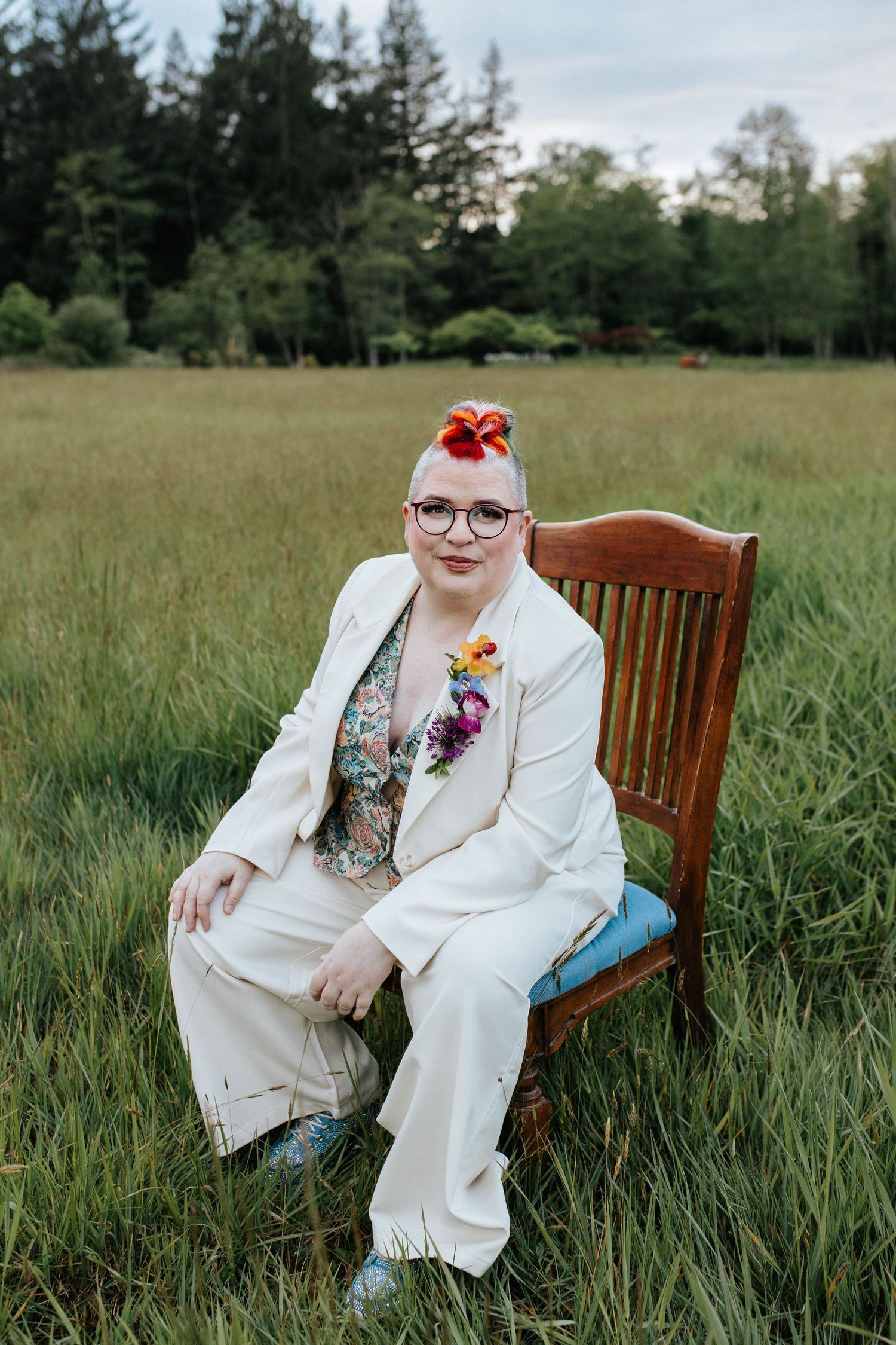 Person in white suit and colorful accessories sits on wooden chair in grassy meadow with evergreen trees behind.
