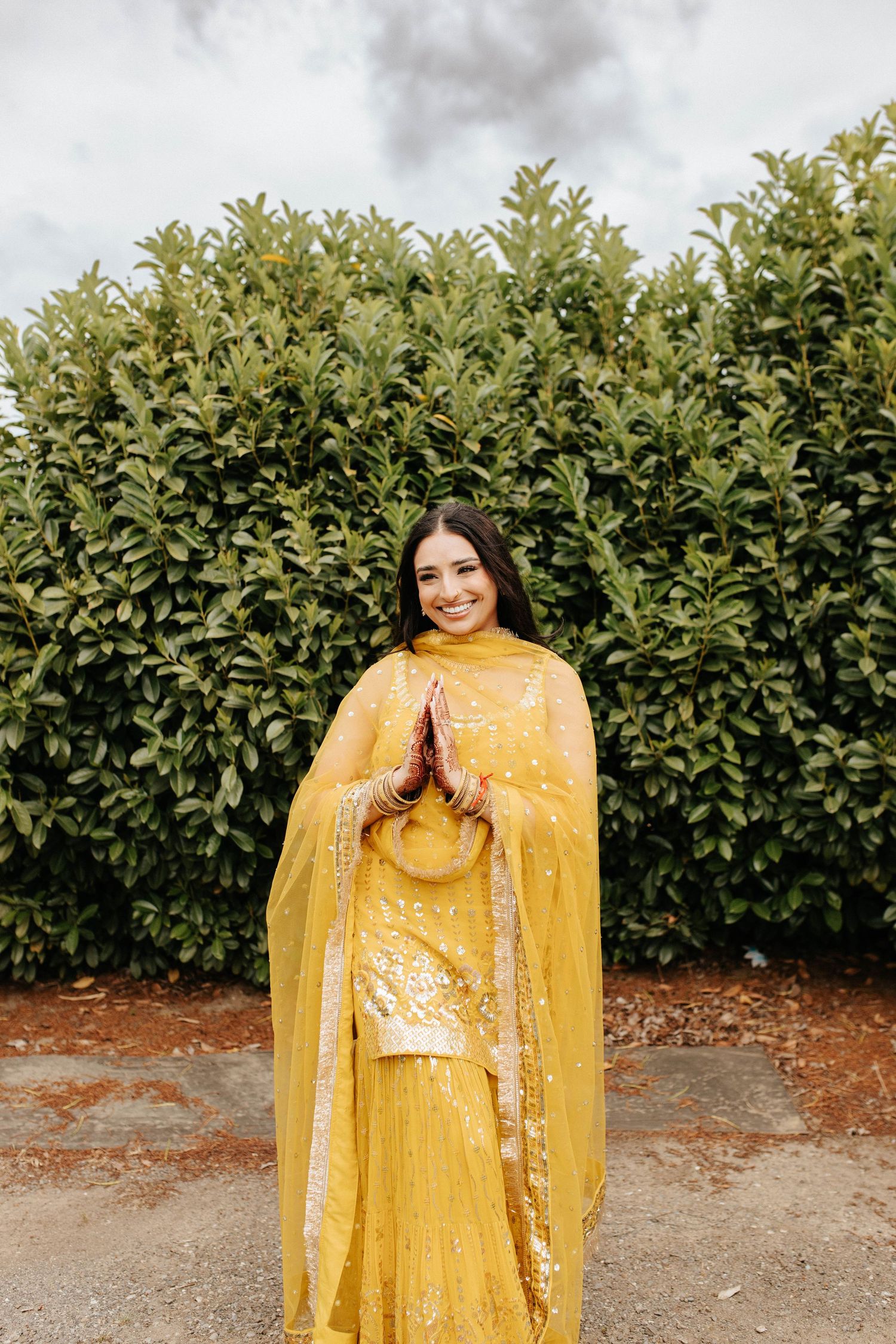 Beautiful yellow traditional Indian lehenga outfit photographed against a green hedge backdrop in outdoor setting.