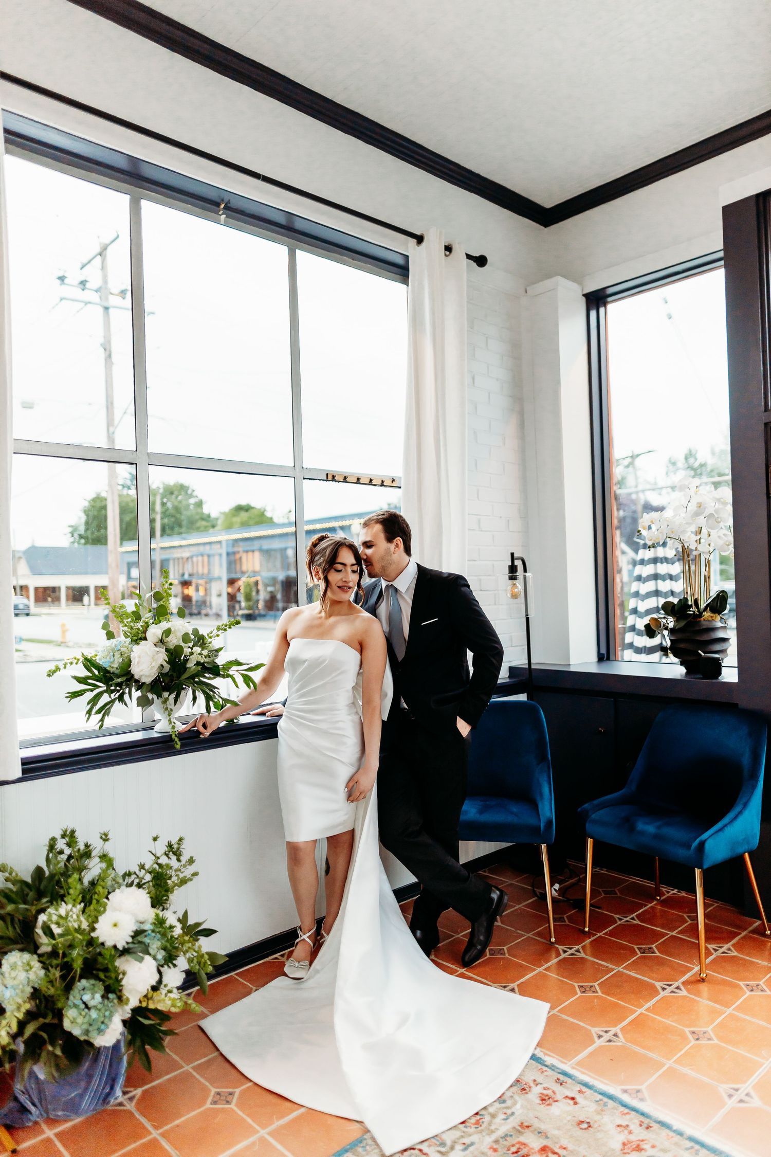 Wedding couple stands together in bright corner room with blue velvet chairs, white floral arrangements and terracotta floors.