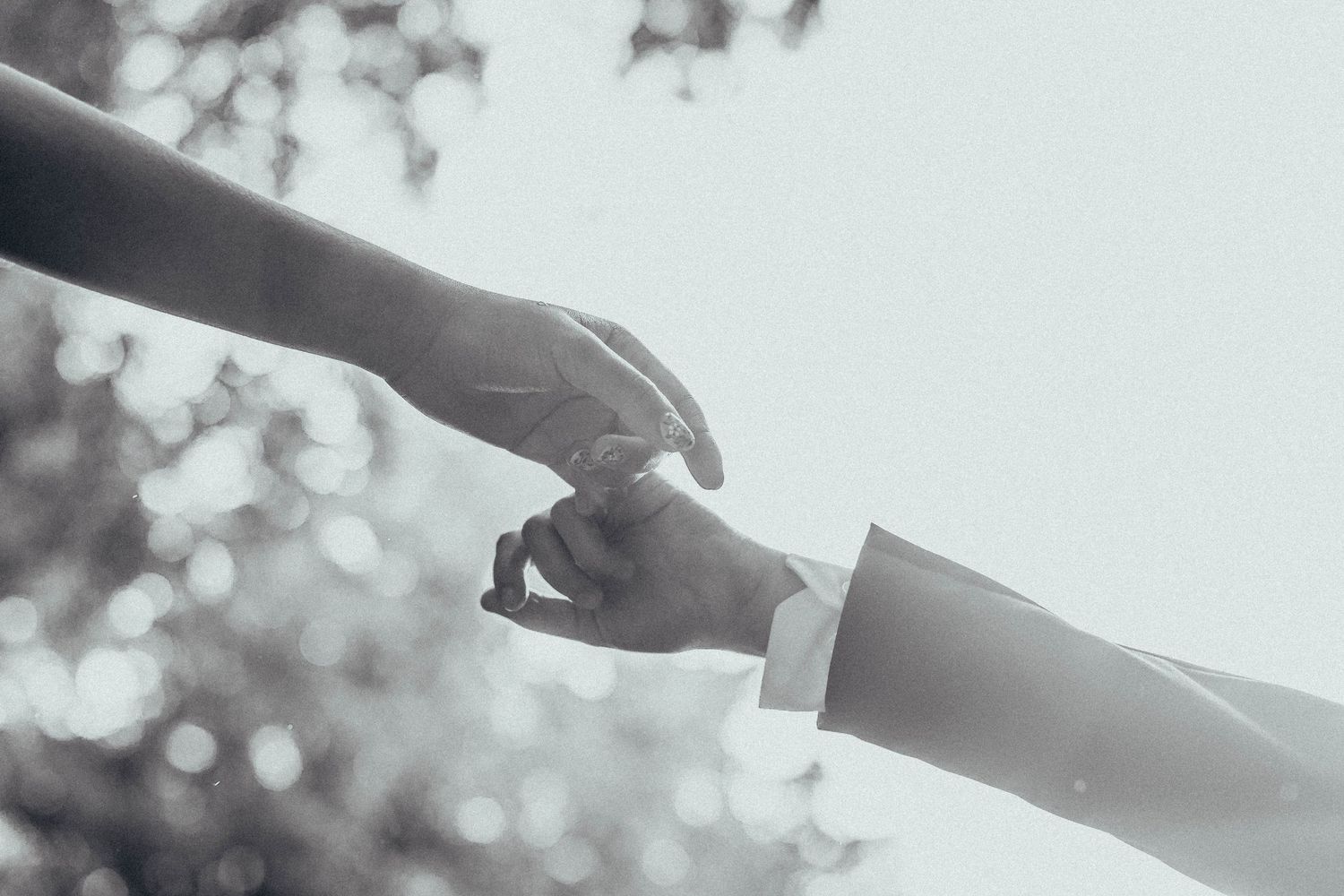 Two hands gently touching fingertips in a romantic black and white wedding photo with soft bokeh background.