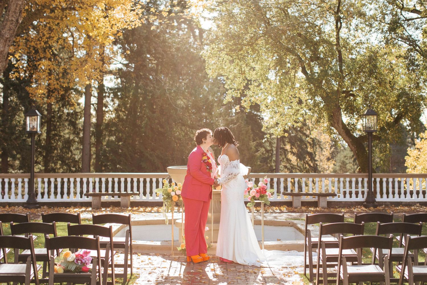 Bride and groom share first kiss at outdoor ceremony surrounded by autumn trees and colorful florals.